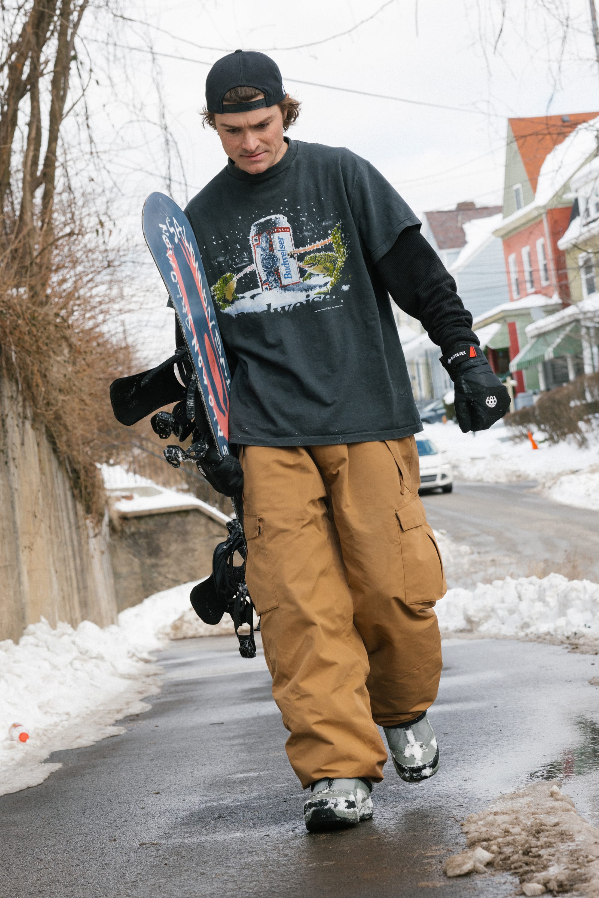 A young man in a black cap, graphic t-shirt, and 686 Men’s Essox™ Cargo Pant walks along a snowy street carrying a snowboard and bindings, with colorful houses in the background.