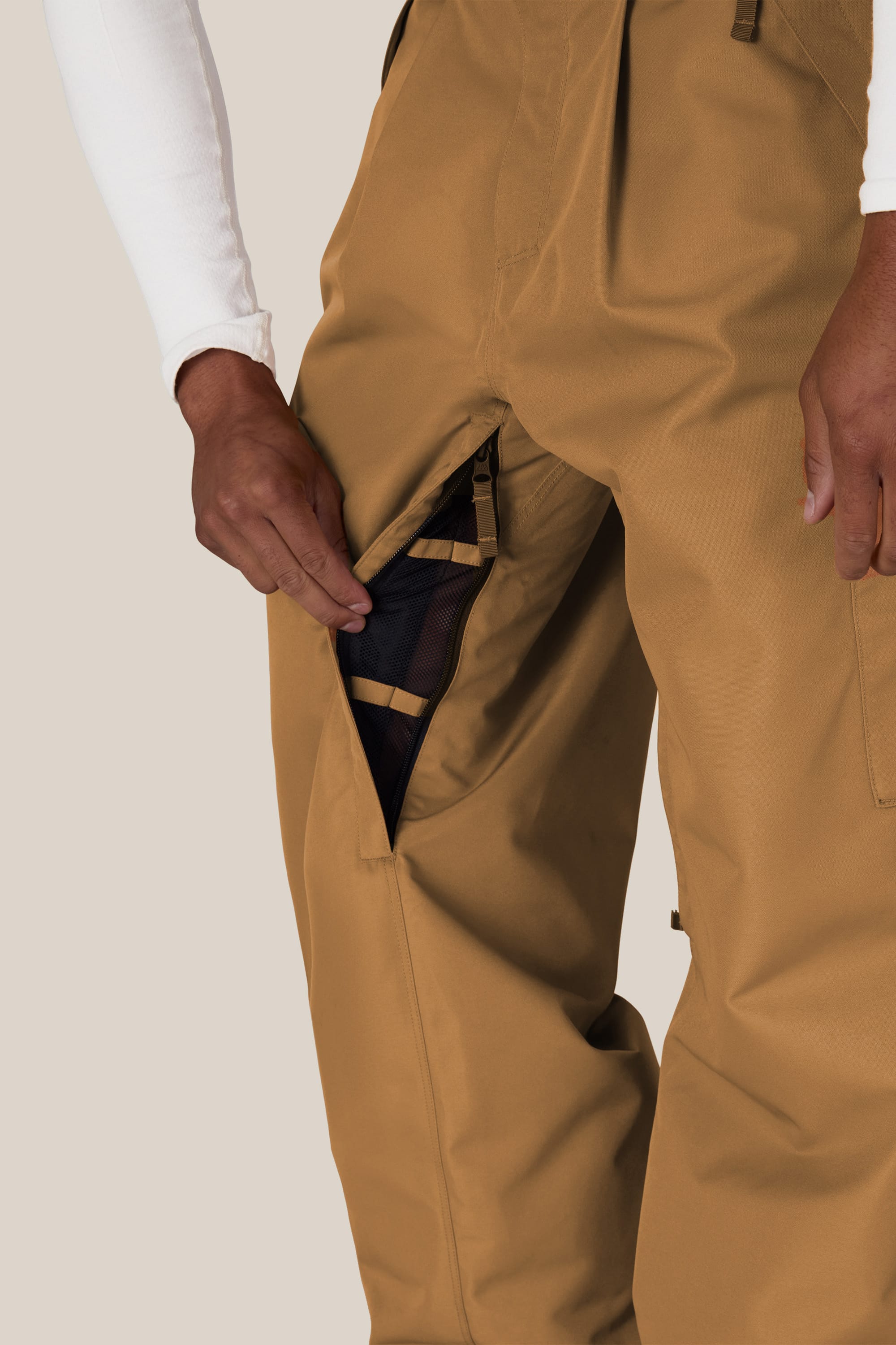 A person wearing the 686 Men’s Essox™ Cargo Pant in tan and a white long-sleeve shirt opens a zippered mesh-lined side vent on their 686 pants, revealing the interior.