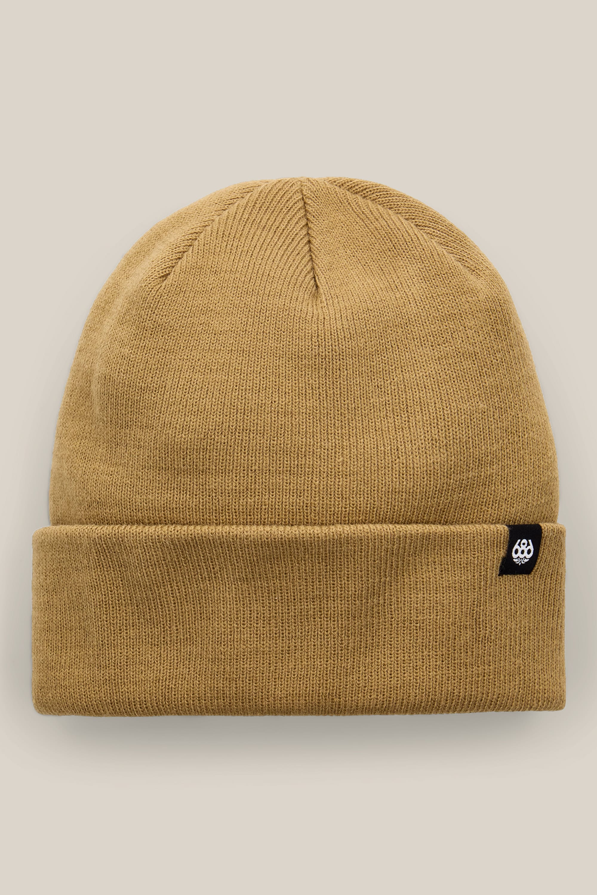 The 686 Standard Roll Up Beanie by 686 is a tan knit hat with a folded brim, accented by a small black tag featuring a white logo on the left side, and is photographed against a light beige background.