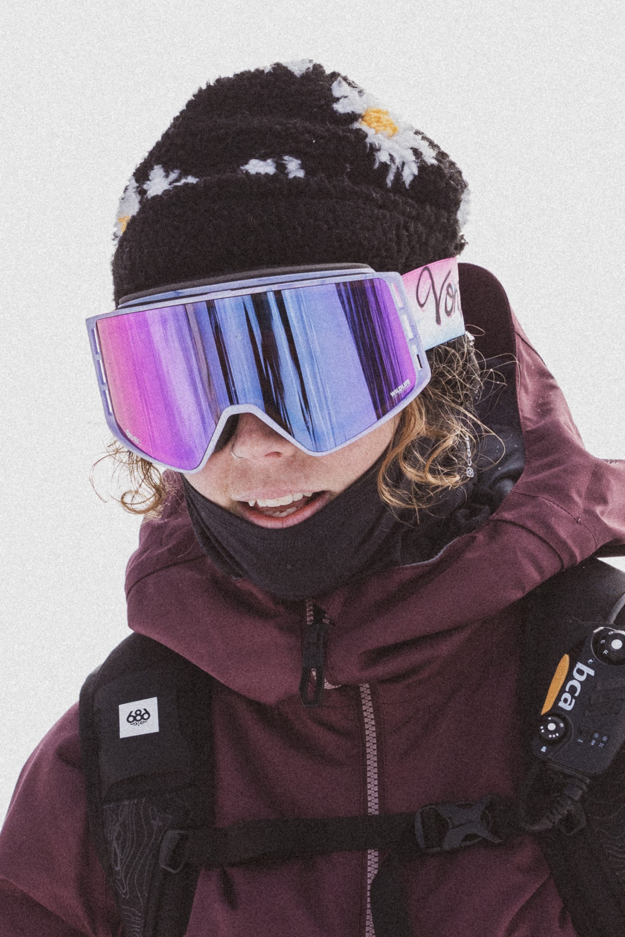 A person in reflective ski goggles, a 686 Dawn Fleece Beanie, and a maroon jacket stands against a snowy backdrop, carrying gear on their back.