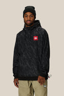 A man wears the 686 Mens Bonded Fleece Pullover Hoody in black with a red and white chest logo, paired with a black beanie and beige pants, standing against a plain light background.