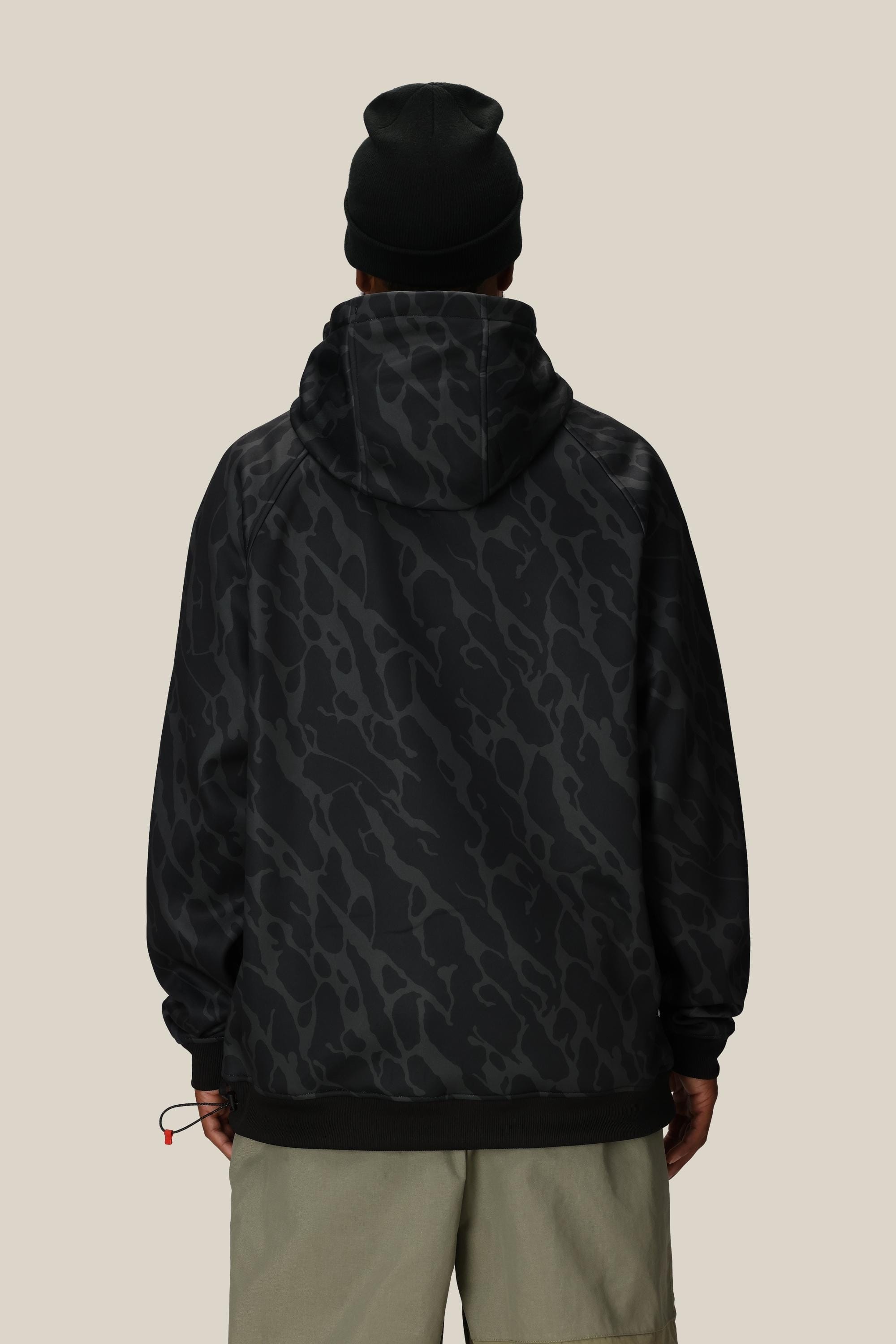 A person stands facing away against a beige background, wearing the 686 Mens Bonded Fleece Pullover Hoody in a black and gray camo pattern, paired with light gray shorts and a black beanie.
