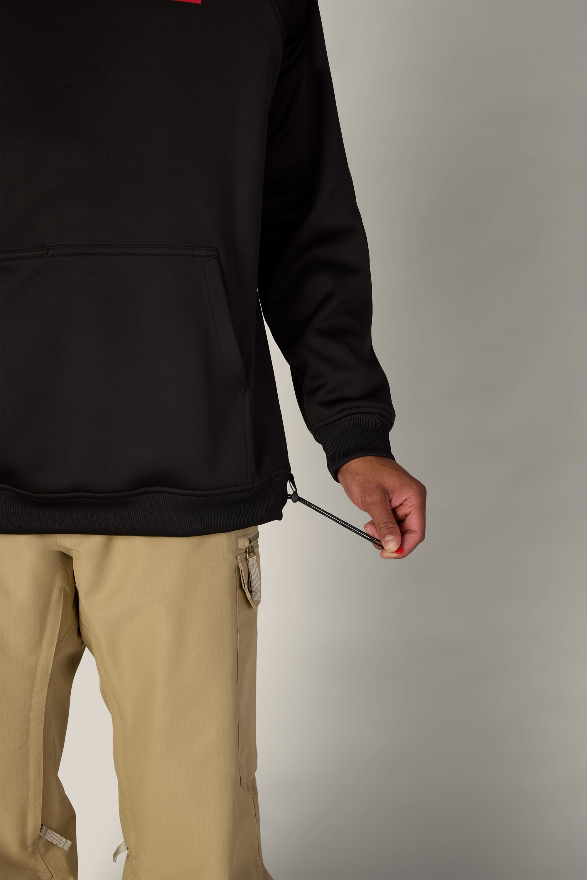 A person dressed in khaki pants and the 686 Men’s Bonded Fleece Pullover Hoody in black adjusts the drawstring at the hem of the hoodie, with only their lower torso and right arm visible against a neutral background.