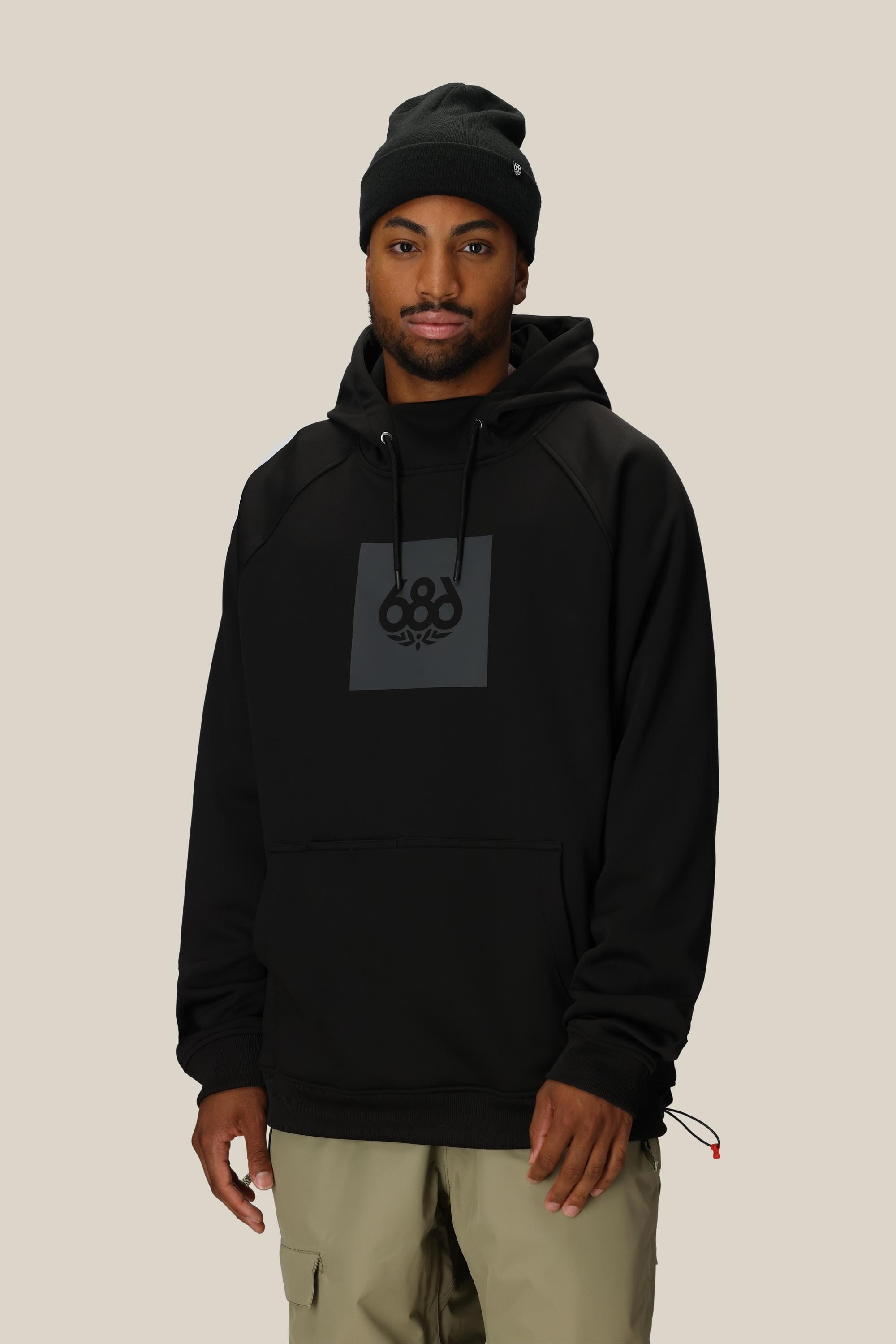 A man wearing a black beanie, light khaki pants, and a 686 Mens Bonded Fleece Pullover Hoody with a gray square logo stands against a plain beige background.