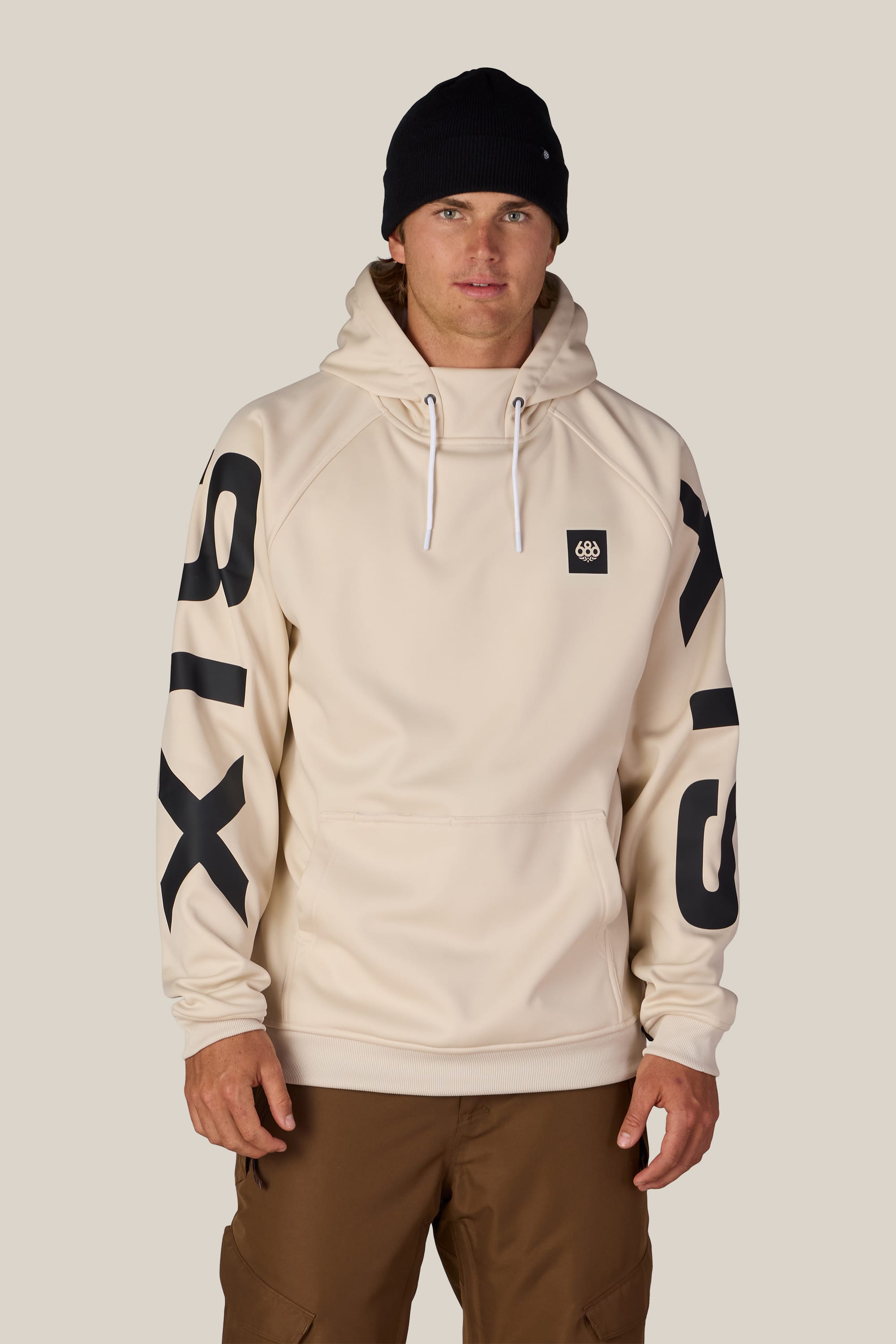 A person wearing the 686 Men’s Bonded Fleece Pullover Hoody in beige, featuring prominent black SIX text on the sleeves, paired with brown pants and a black beanie, standing against a plain light background.