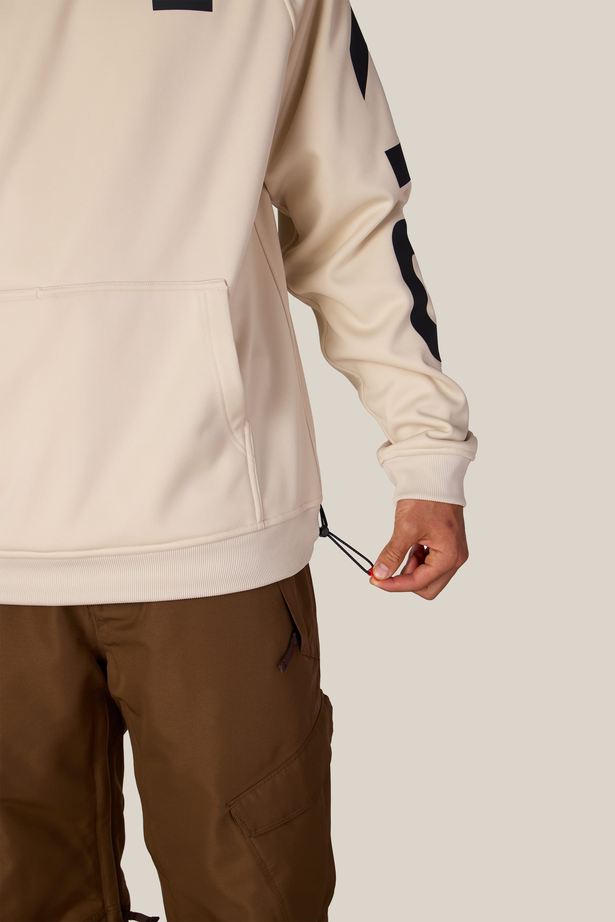 A person wearing the 686 Men’s Bonded Fleece Pullover Hoody in beige and brown pants adjusts the side hem drawstring of the hoodie. Only the lower half of their body and one hand are visible against a plain background.