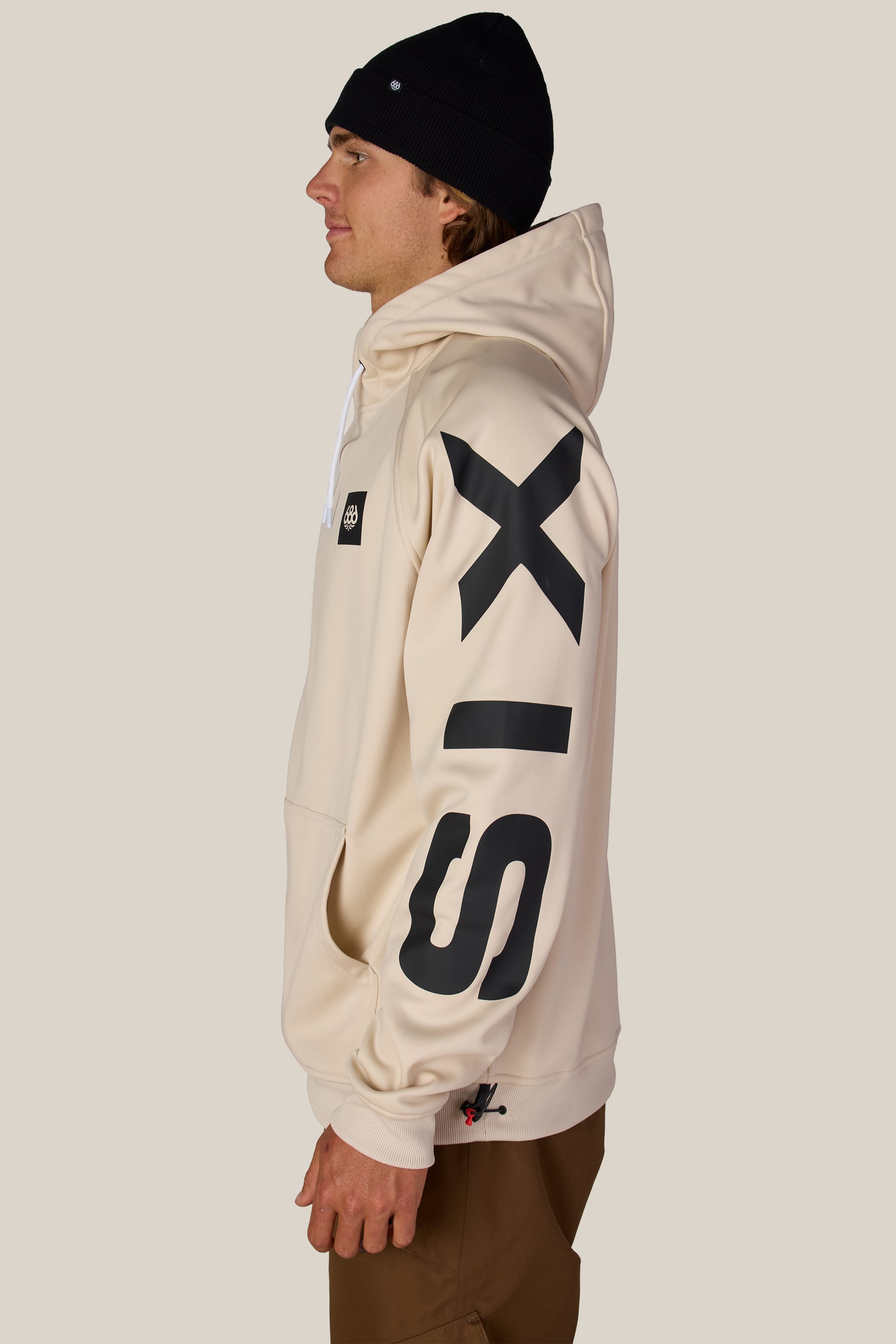 A person stands in profile against a plain light background, wearing the 686 Men’s Bonded Fleece Pullover Hoody in cream with “SIX” in black letters on the sleeve, brown pants, and a black beanie.