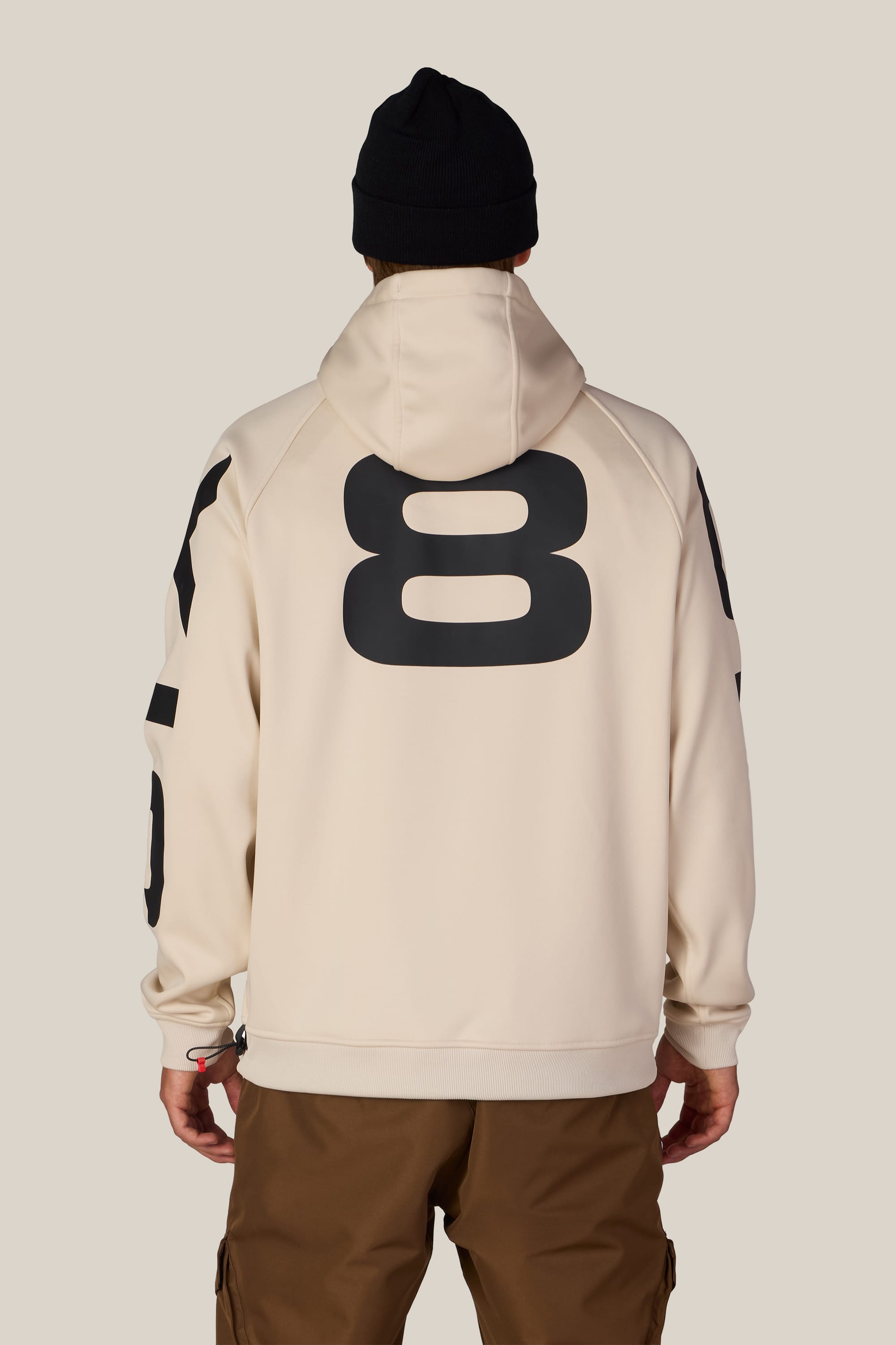 A person is shown from behind against a plain background wearing the 686 Men’s Bonded Fleece Pullover Hoody in beige, featuring a large black number 8 on the back, paired with brown pants and a black beanie.