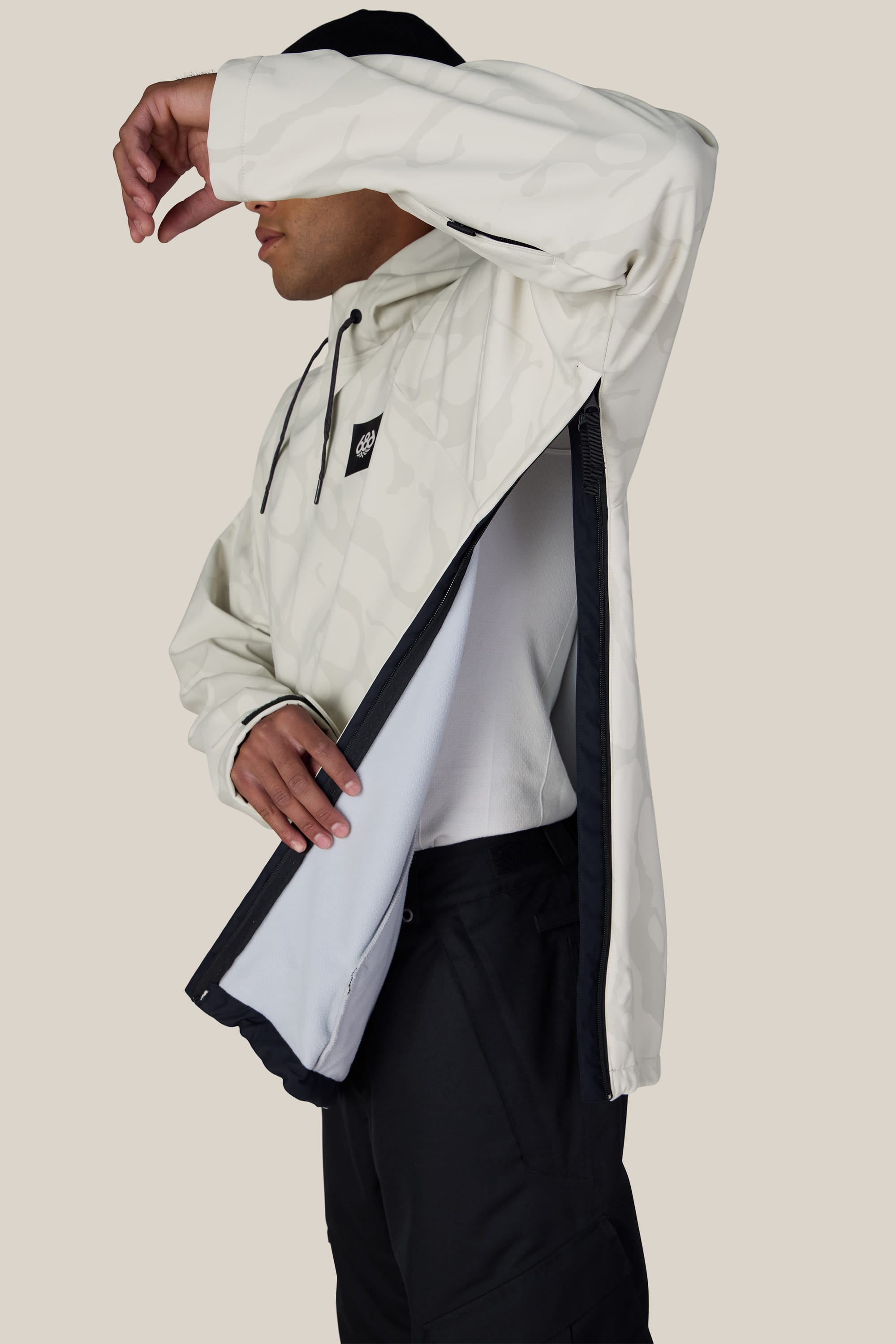 A man models the 686 Men’s Waterproof Hyperchromic Pullover Hoody in a light color with black pants, lifting one arm to reveal the large side zipper opening and the inner lining of the hoody against a plain background.