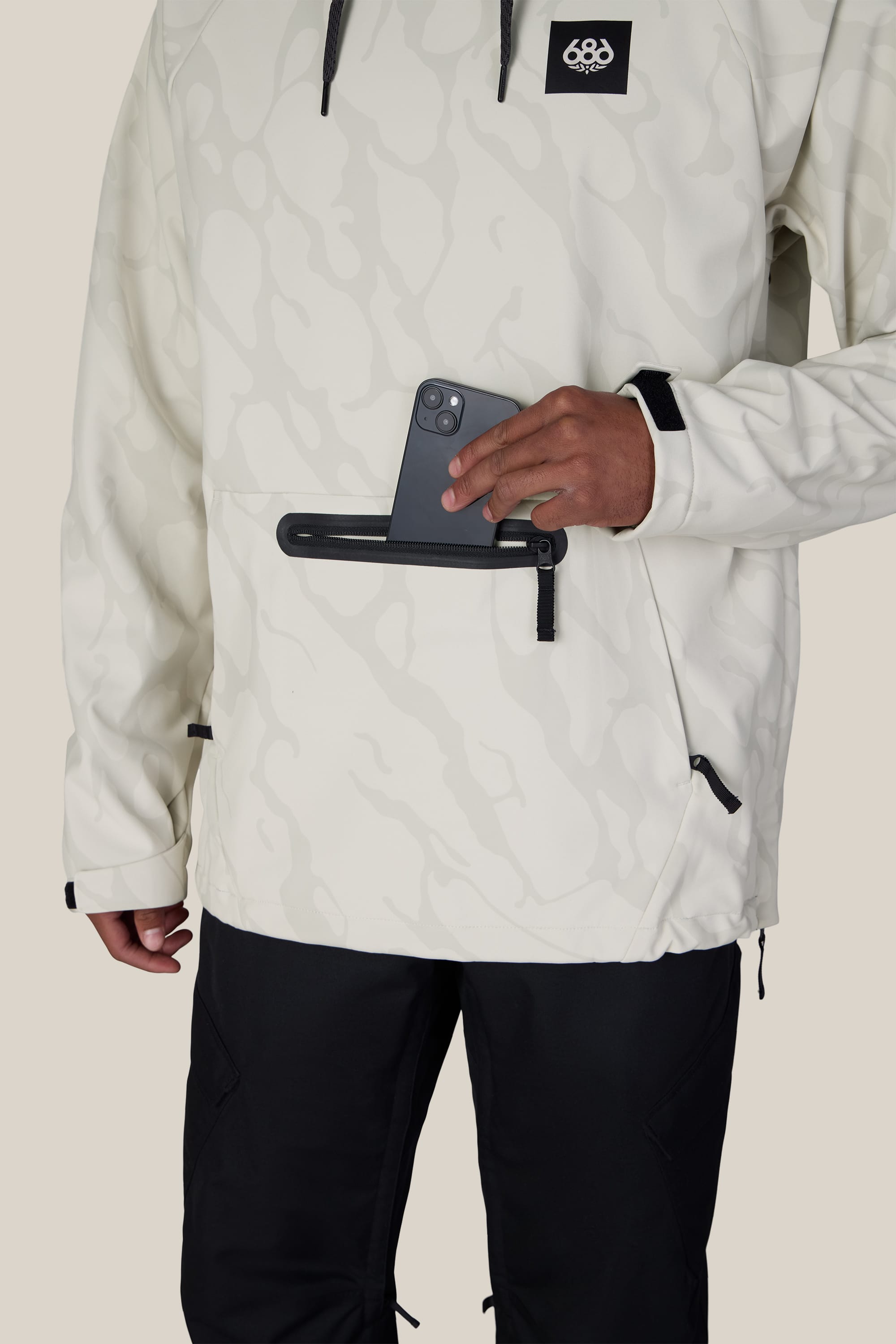 A person wearing the 686 Men’s Waterproof Hyperchromic Pullover Hoody and black pants puts a smartphone into a horizontal zippered pocket on the front of the hoodie.