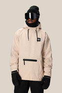 A person wears the 686 Mens Waterproof Hyperchromic Pullover Hoody—a light beige, 10k waterproof jacket with a front zipper pocket—paired with black gloves, pants, beanie, balaclava, and large black ski goggles against a plain background.