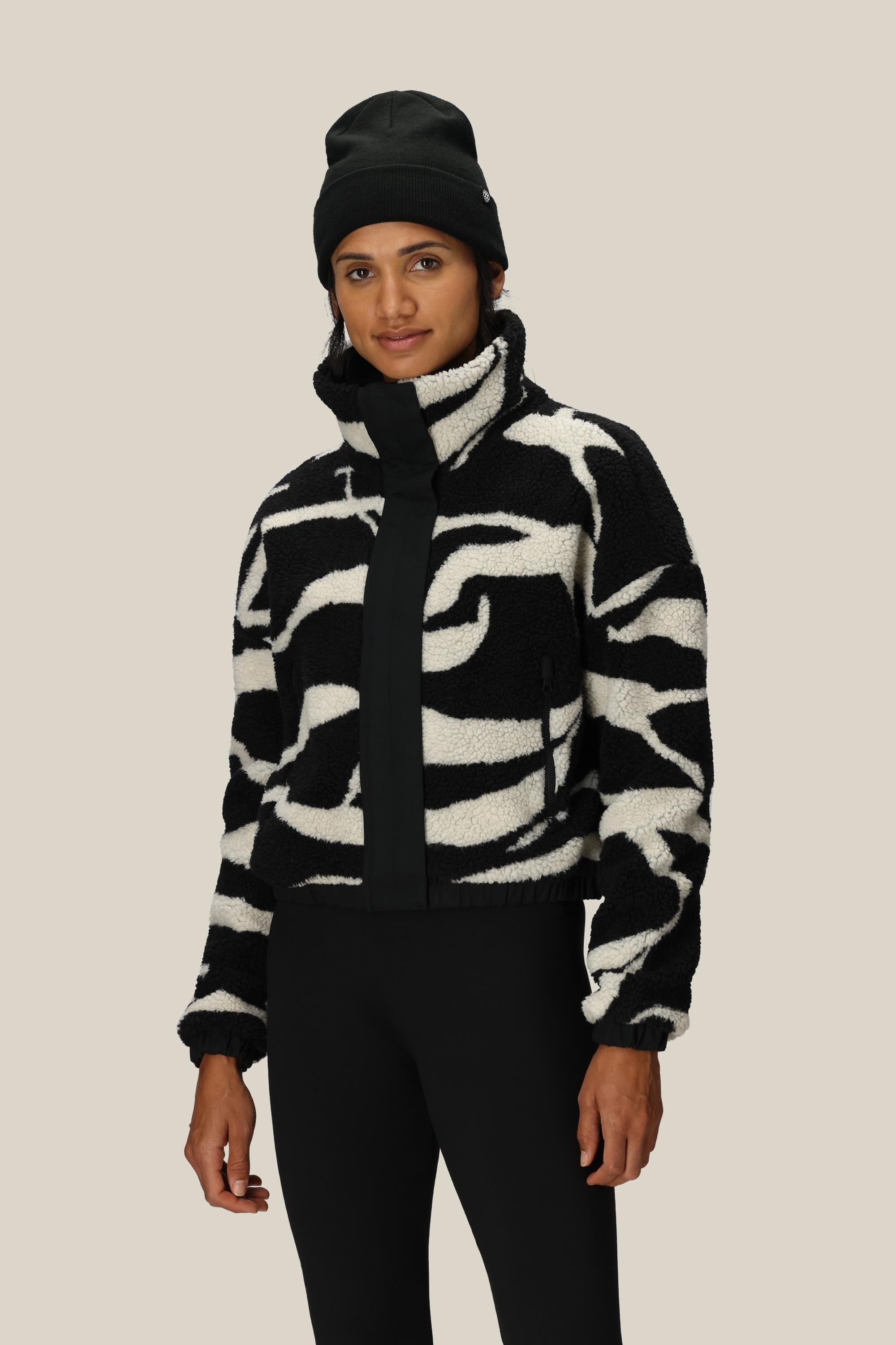 A woman wearing the 686 Women’s Outpost Sherpa Jacket by 686—featuring a black-and-white abstract pattern—paired with a black beanie and black pants, stands against a plain beige background.