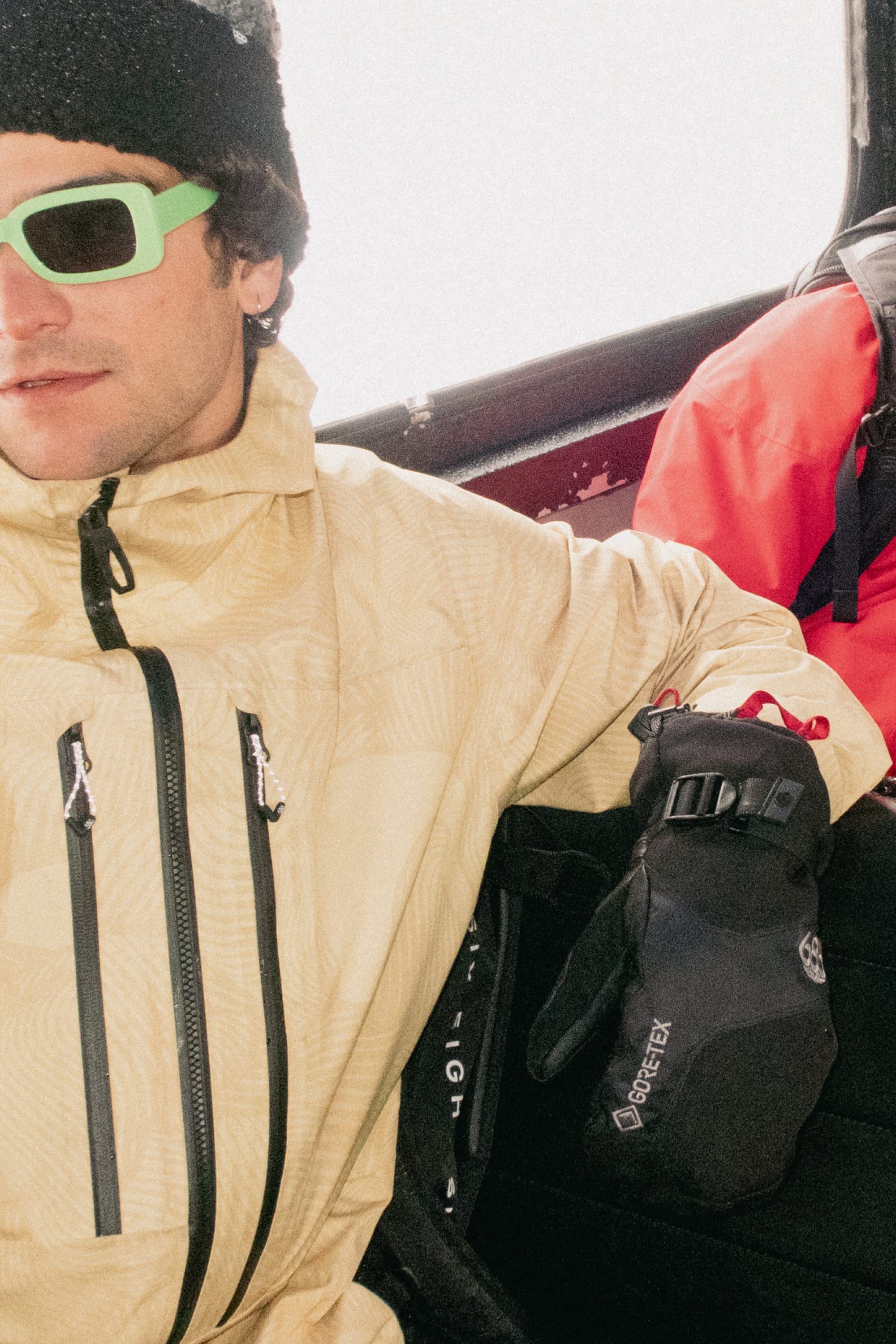 Wearing a light yellow jacket, green sunglasses, and black beanie, a person sits with the 686 Mens GORE-TEX Leather Linear Mitt by 686 on their lap. A red backpack is beside them as they ride what appears to be a ski lift or gondola.
