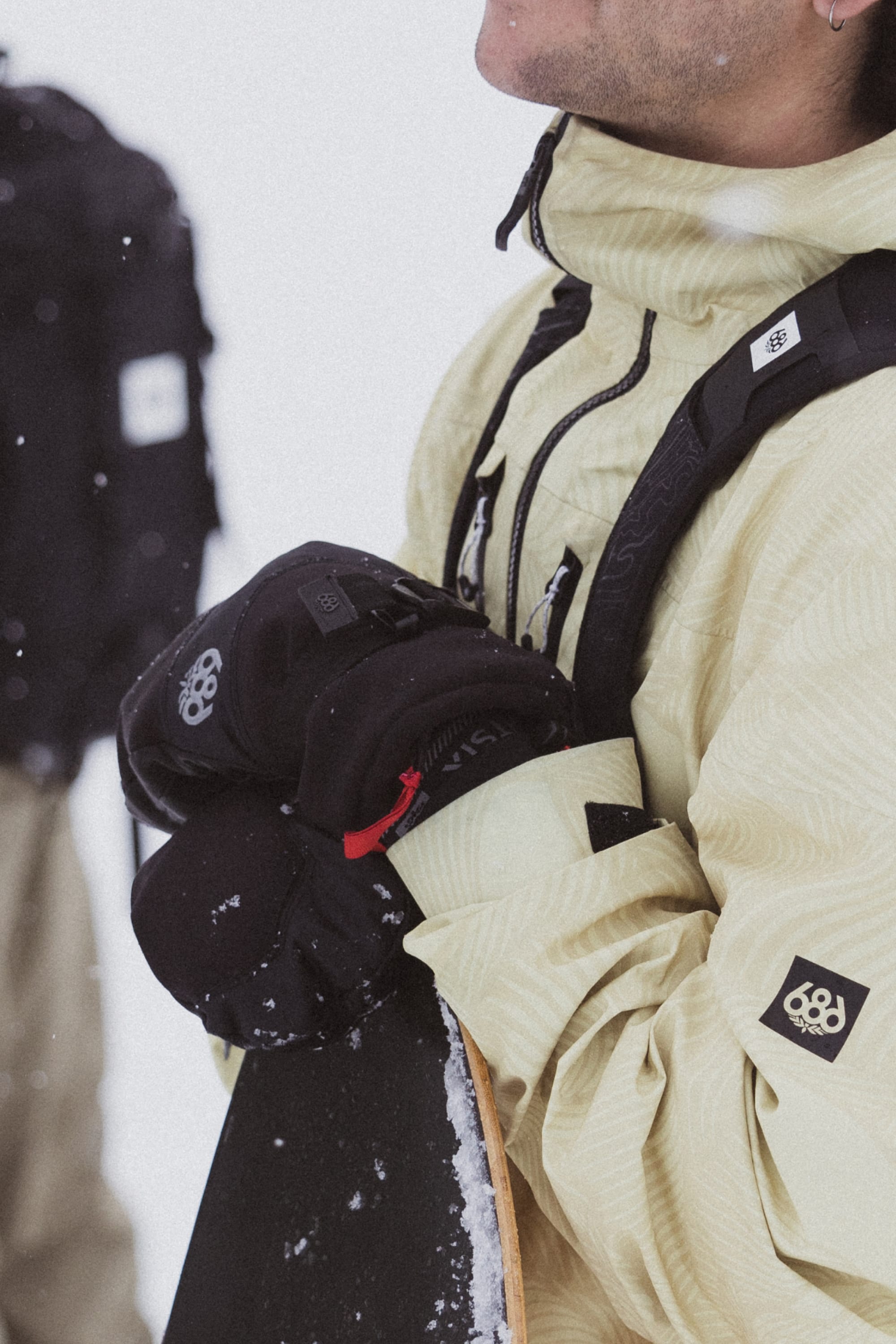 Wearing a light-colored winter jacket and 686 Mens GORE-TEX Leather Linear Mitt, someone holds a snowboard upright in a snowy outdoor setting with falling snowflakes; another person is partially visible in the background.