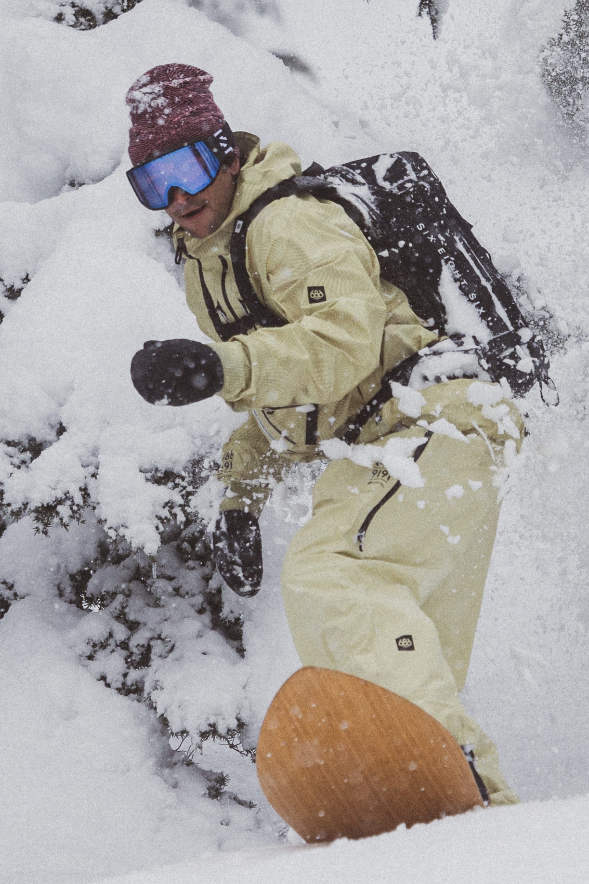 A snowboarder in beige gear, maroon beanie, blue goggles, and a backpack rides through fresh snow with snow-covered trees, hands protected by 686 Mens GORE-TEX Leather Linear Mitts.