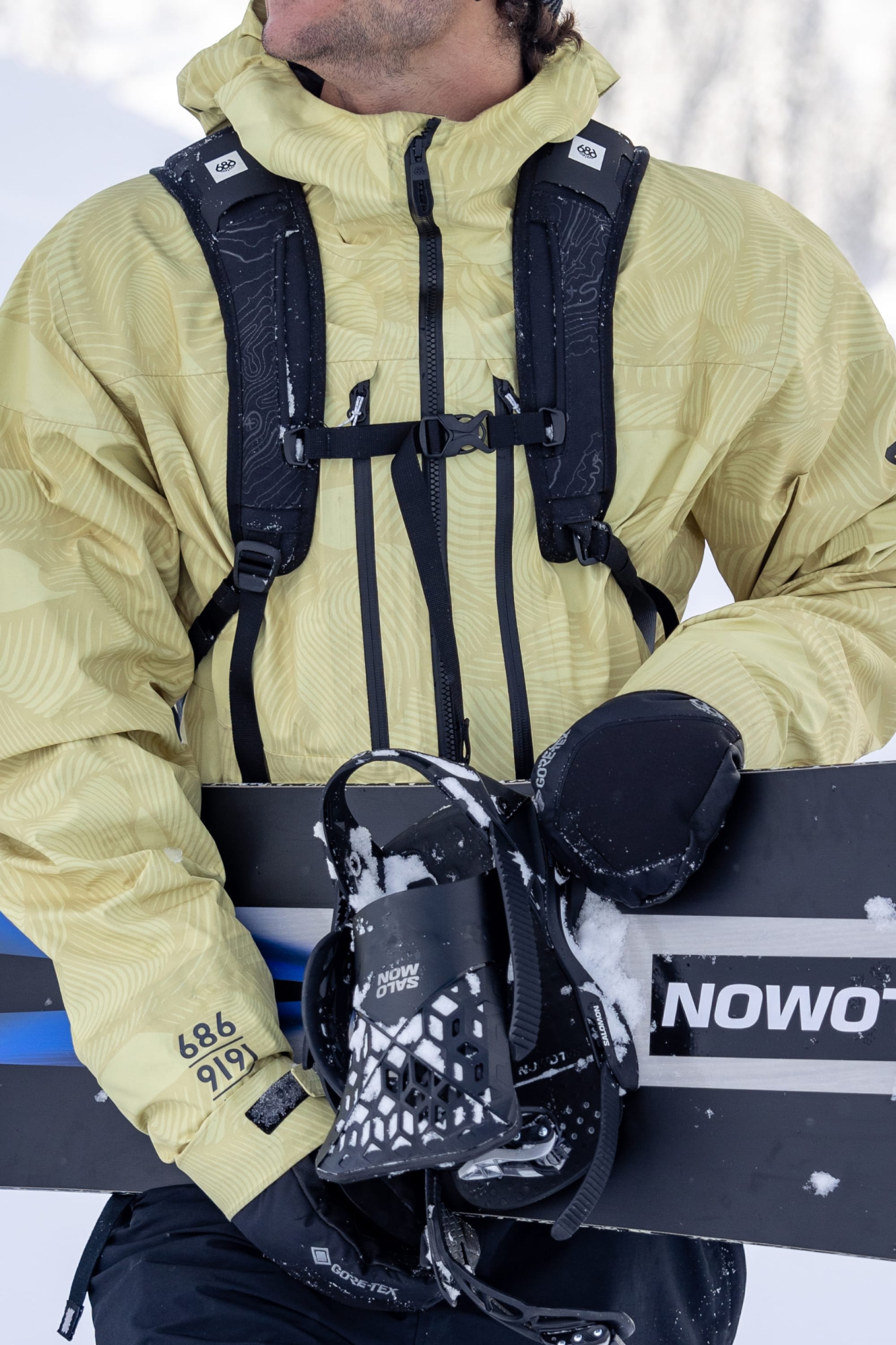 Wearing a yellow jacket with black straps and 686 Mens GORE-TEX Leather Linear Mitts, a person holds a snowboard marked NOWO and a black helmet in snowy outdoor conditions.