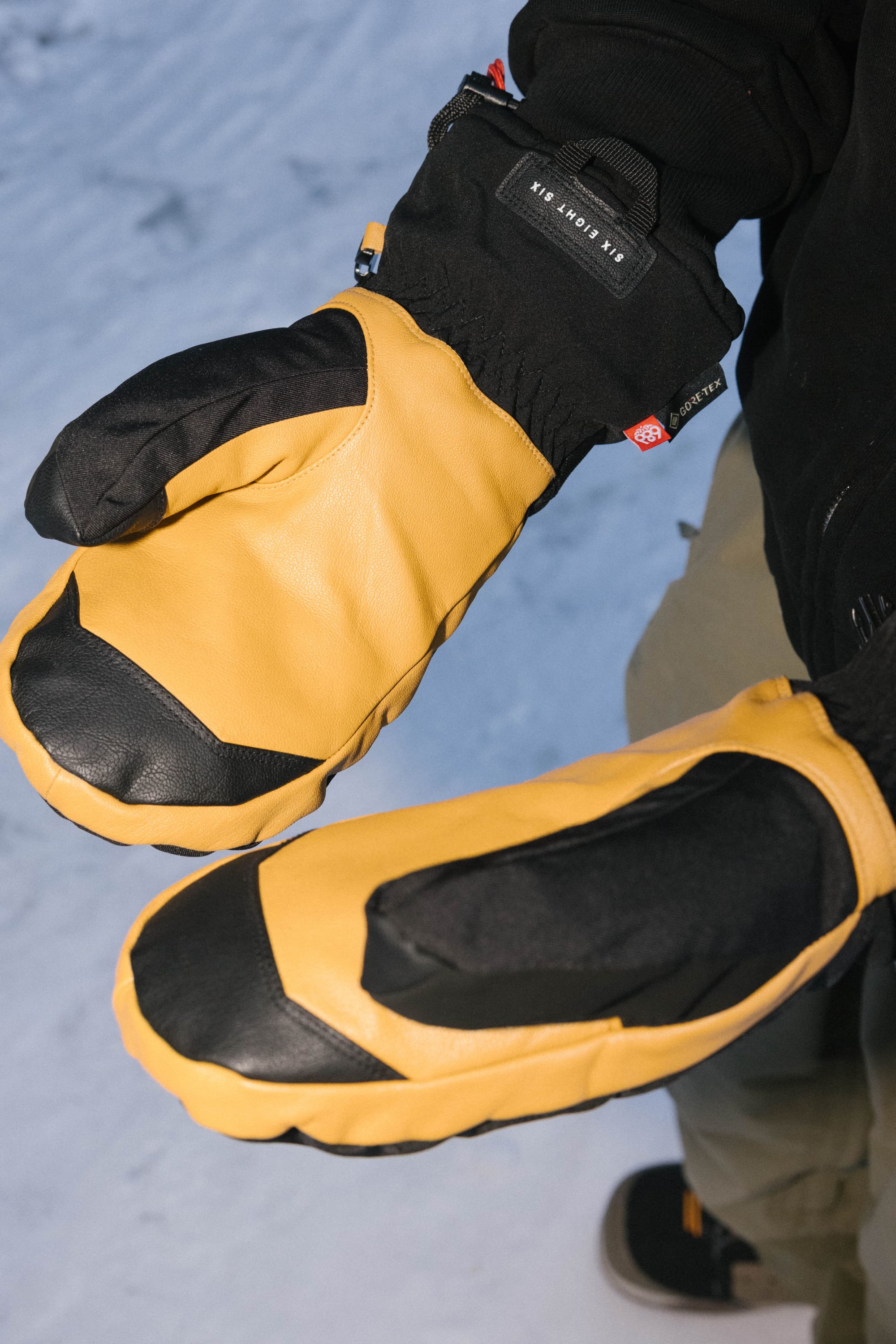 A person wearing 686 Mens GORE-TEX Leather Linear Mitts in yellow and black stands on snow, raising both hands to show the leather-palmed mittens clearly against a dark jacket and greenish pants.