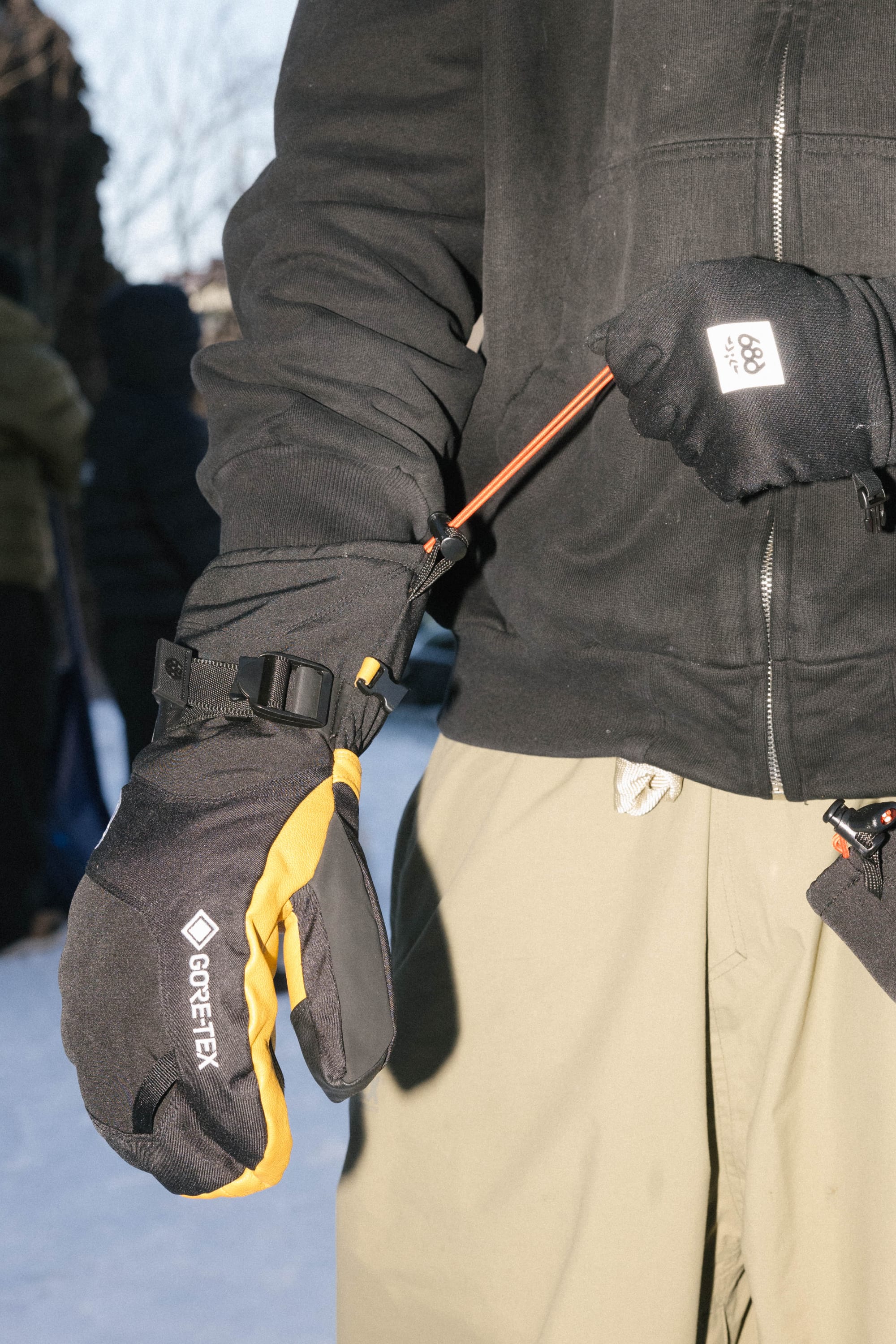A person in a black jacket and tan pants tightens the strap on a 686 Mens GORE-TEX Leather Linear Mitt with a ski pole. The black and yellow mitt from 686 features a leather palm. Snow and other people can be seen in the background.
