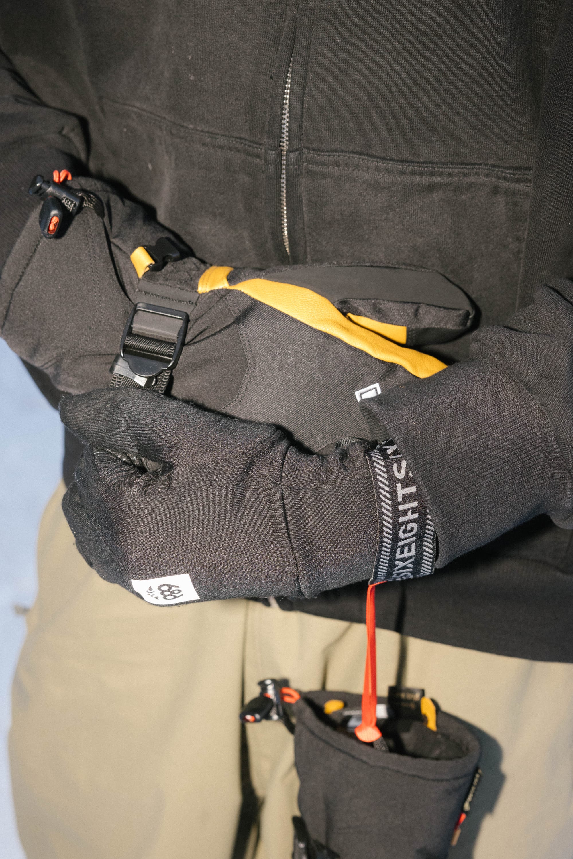 Close-up of a person wearing 686 Mens GORE-TEX Leather Linear Mitts in black, adjusting a black and yellow strap with a buckle. Light brown pants and another strap featuring a red cord are partially visible. Brand: 686.