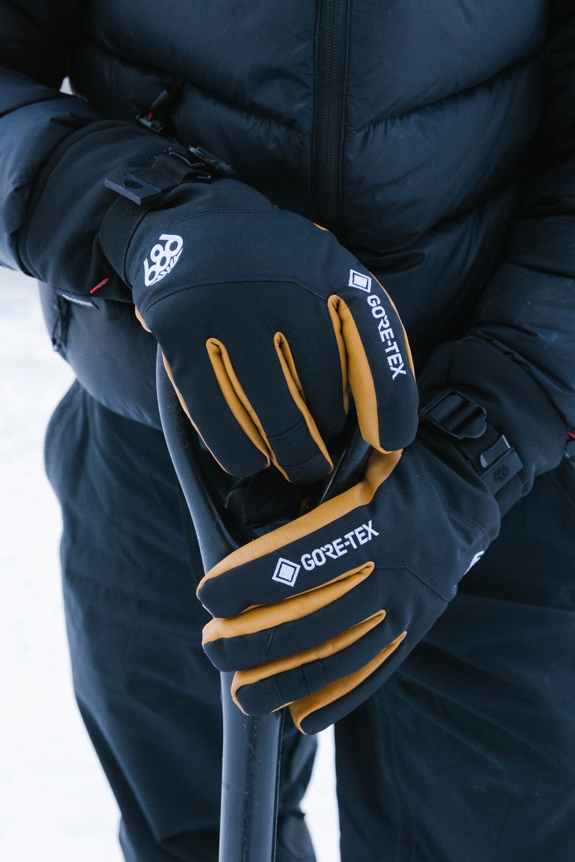 A person wearing 686 Men’s GORE-TEX® Leather Linear™ Gloves in black and brown, along with a black puffer jacket, is holding a snow shovel outdoors. Only the gloved hands featuring the 686 brand, part of the shovel handle, and jacket are visible.
