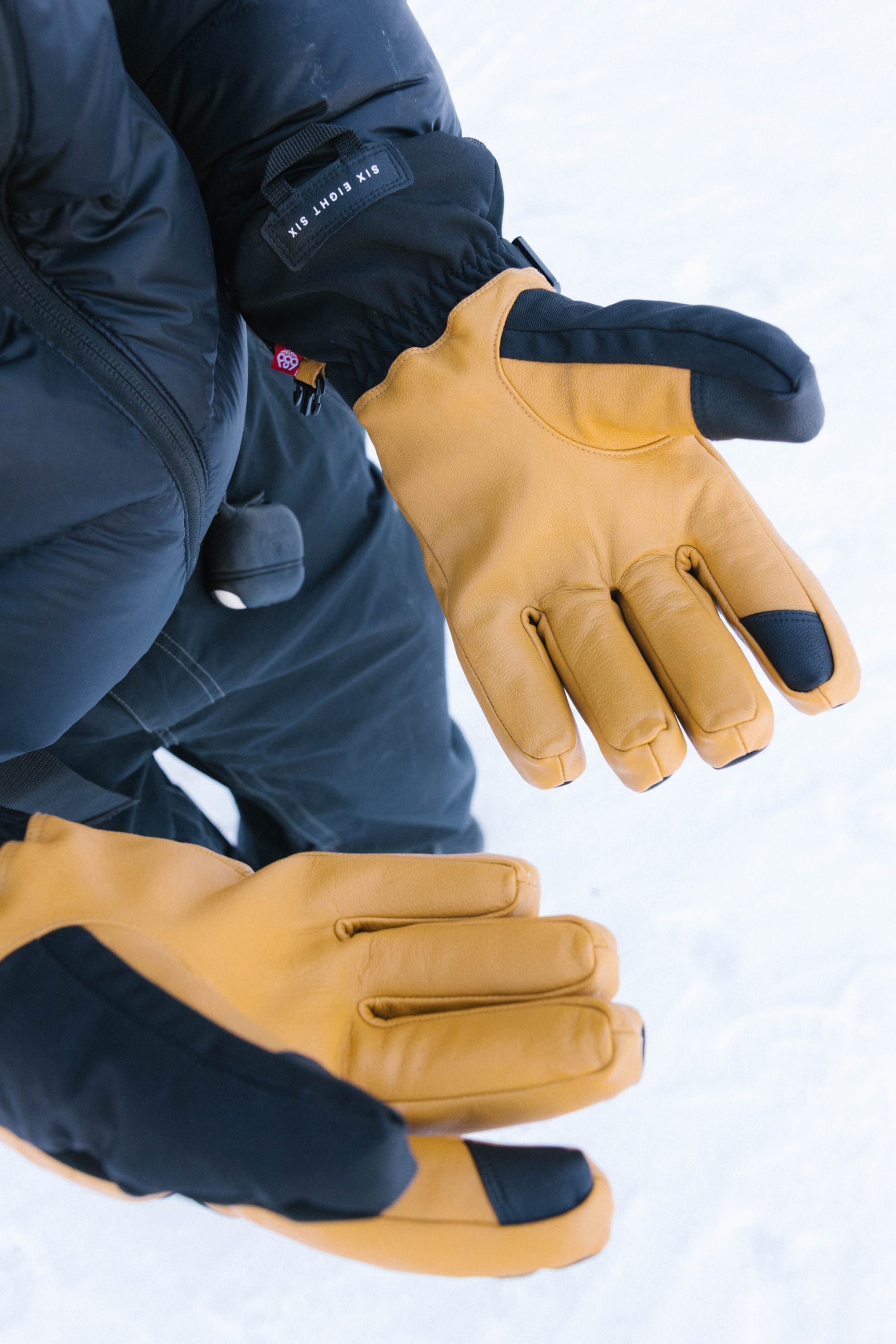 Wearing the 686 Men’s GORE-TEX® Leather Linear™ Gloves in black and yellow, a person holds their hands open over the snow, dressed in a dark winter jacket and pants.