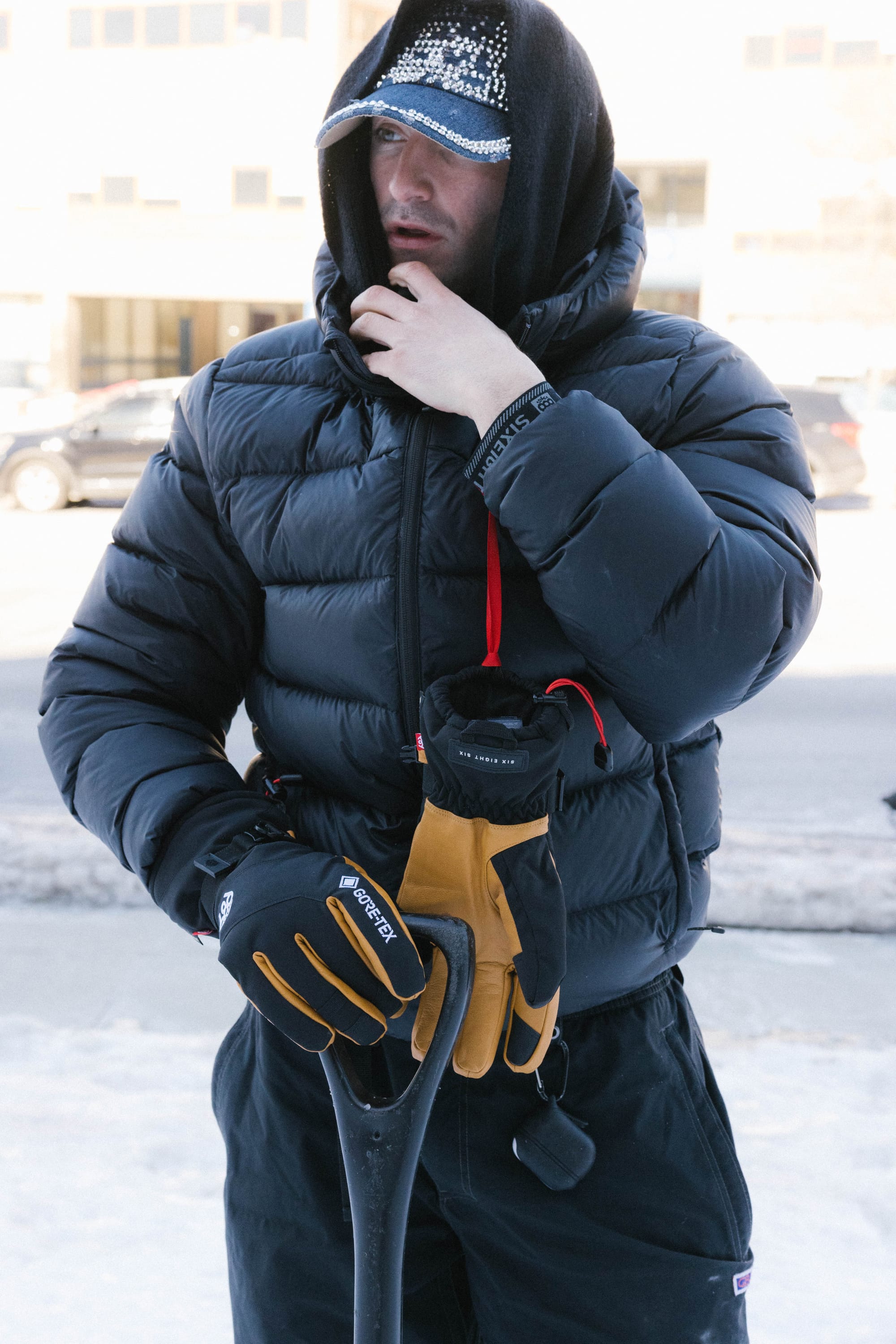 A person in a black puffer jacket and cap stands outside on a snowy day, holding a shovel while wearing 686 Men’s GORE-TEX® Leather Linear™ Gloves for warmth, their hood up and face partly covered.