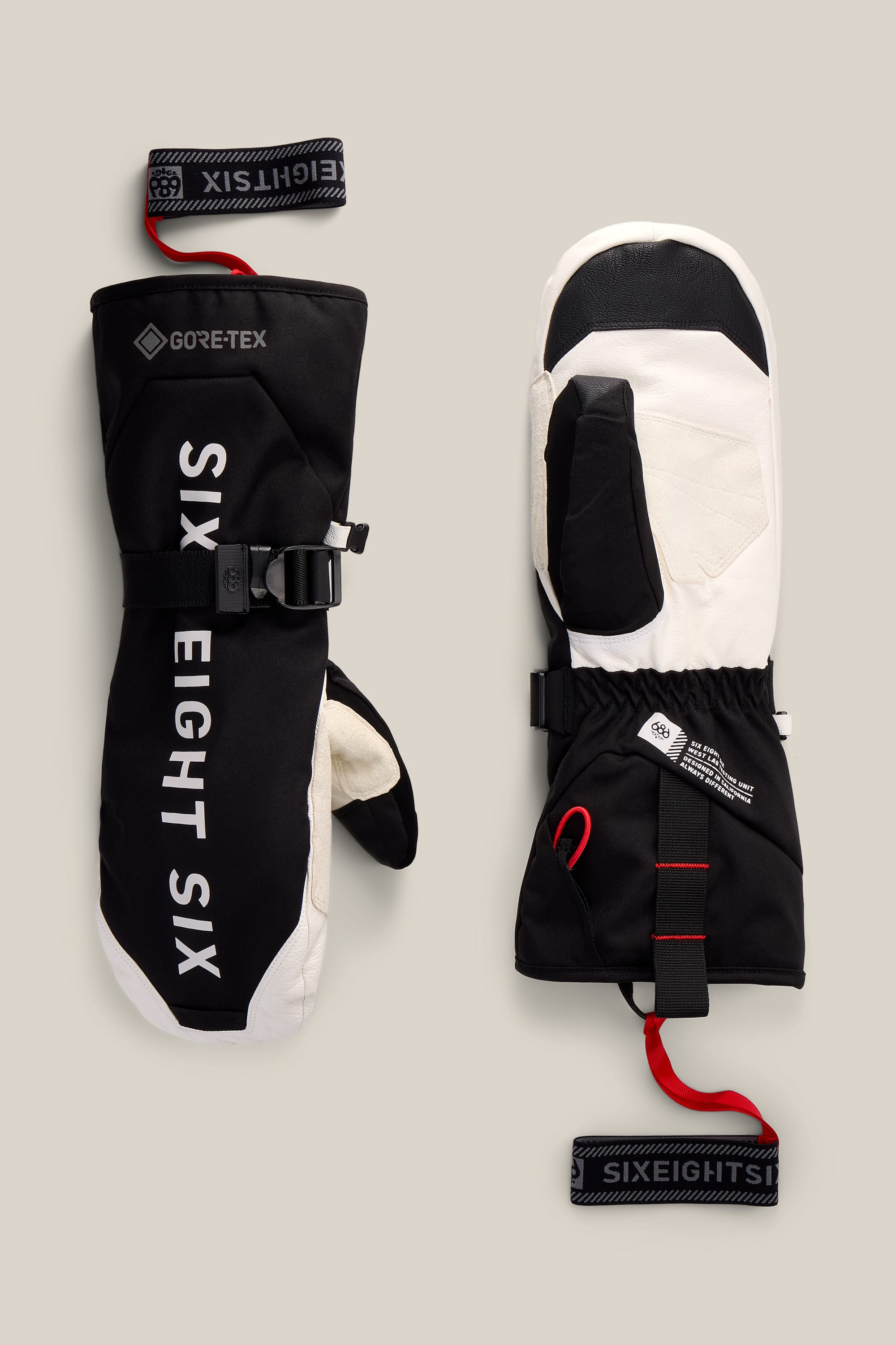 A pair of black and white 686 Men’s GORE-TEX® Leather Deep Days™ Mitts with adjustable straps and loops, displayed on a beige background.