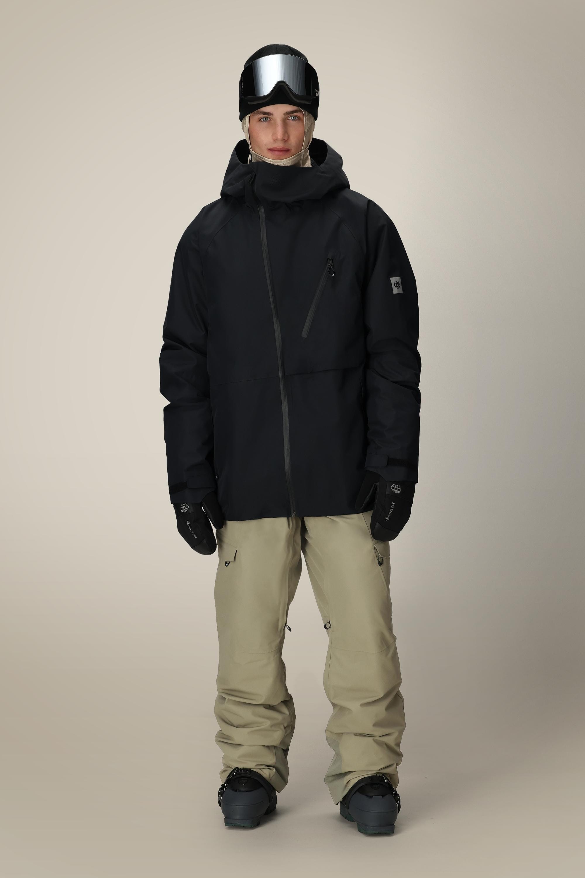 A person wearing the 686 Men’s Hydra™ Thermagraph® Jacket by 686, along with tan pants, black gloves, black boots, a black helmet, and reflective ski goggles stands facing forward against a plain light background.