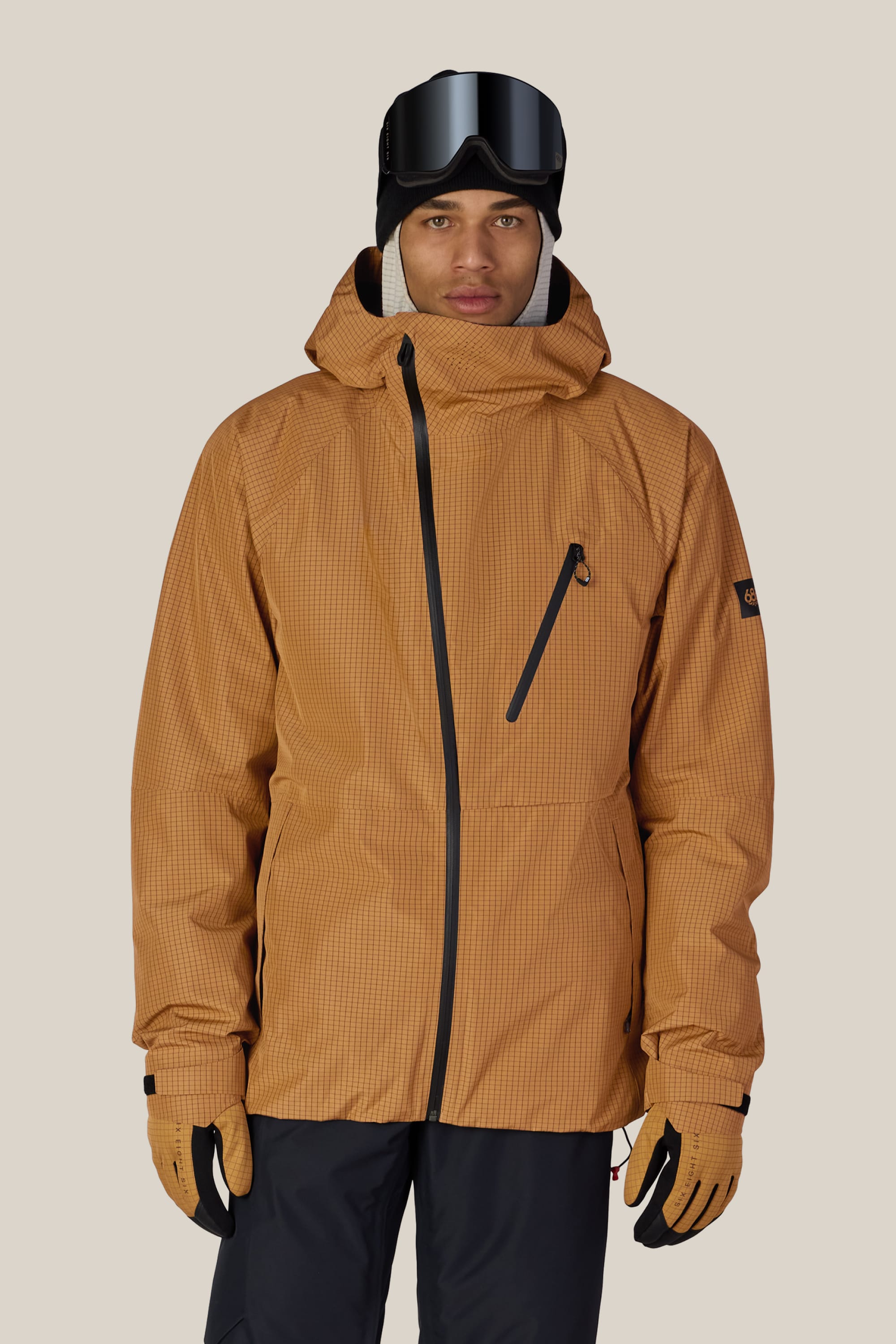 A person wearing the 686 Men’s Hydra™ Thermagraph® Jacket by 686, along with black ski goggles, black pants, tan gloves, and a black beanie stands facing forward against a plain light background.