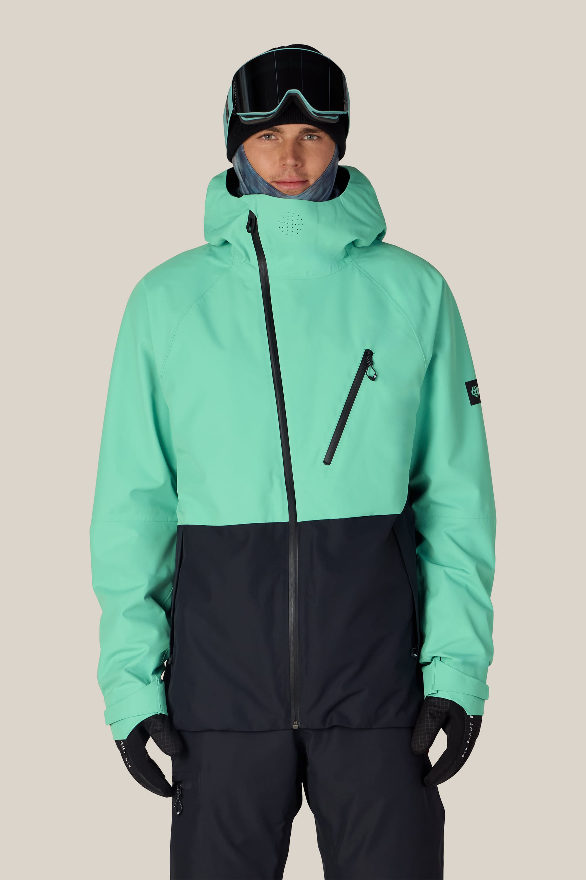 A person wearing the 686 Men’s Hydra™ Thermagraph® Jacket in teal and black, paired with black ski gloves, black snow pants, ski goggles, and a blue neck gaiter, stands against a plain light background.