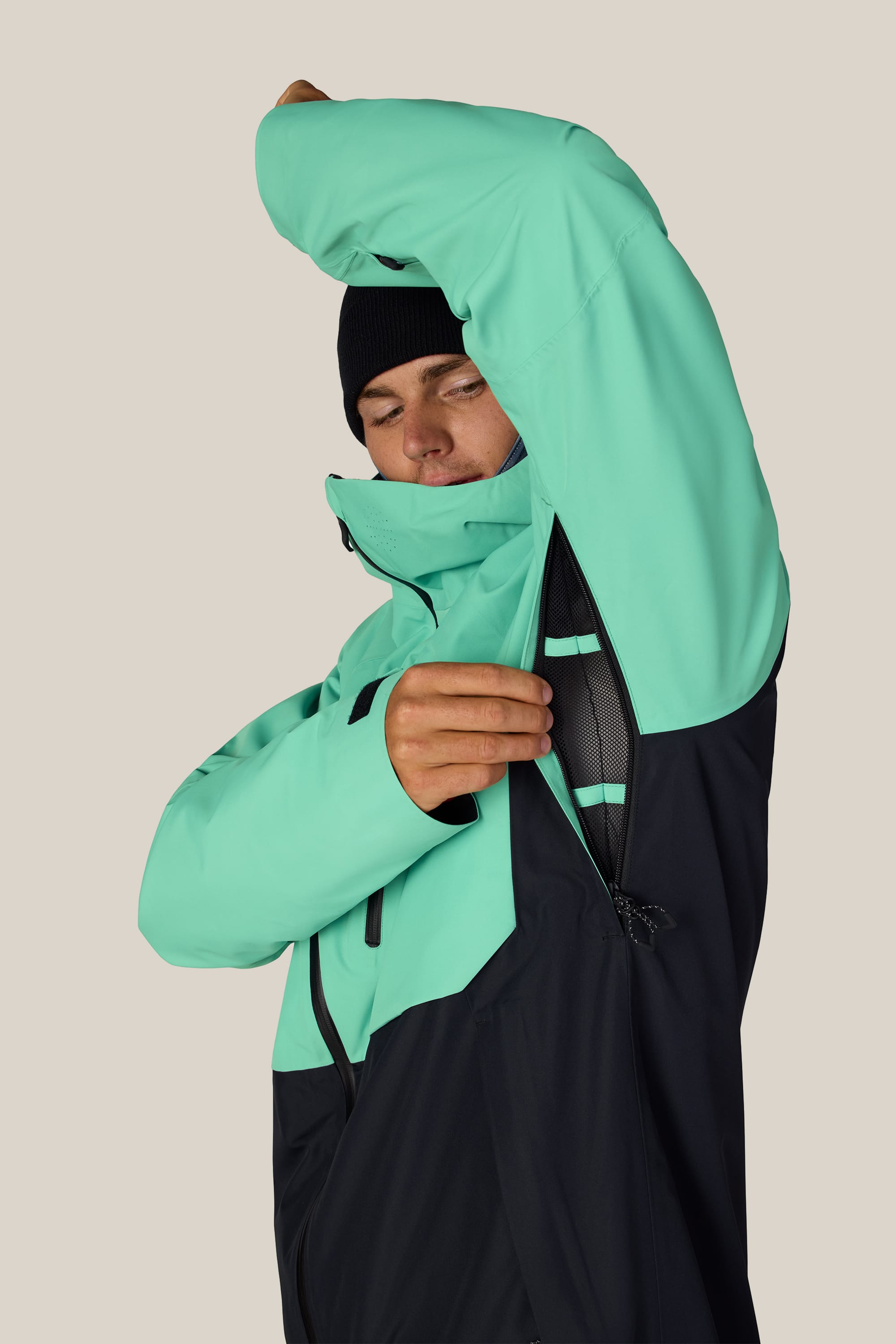 A person wearing the 686 Men’s Hydra™ Thermagraph® Jacket in turquoise and black unzips the underarm vent. They have a black beanie on and one arm raised, looking down at the opened vent. The background is plain and light-colored.