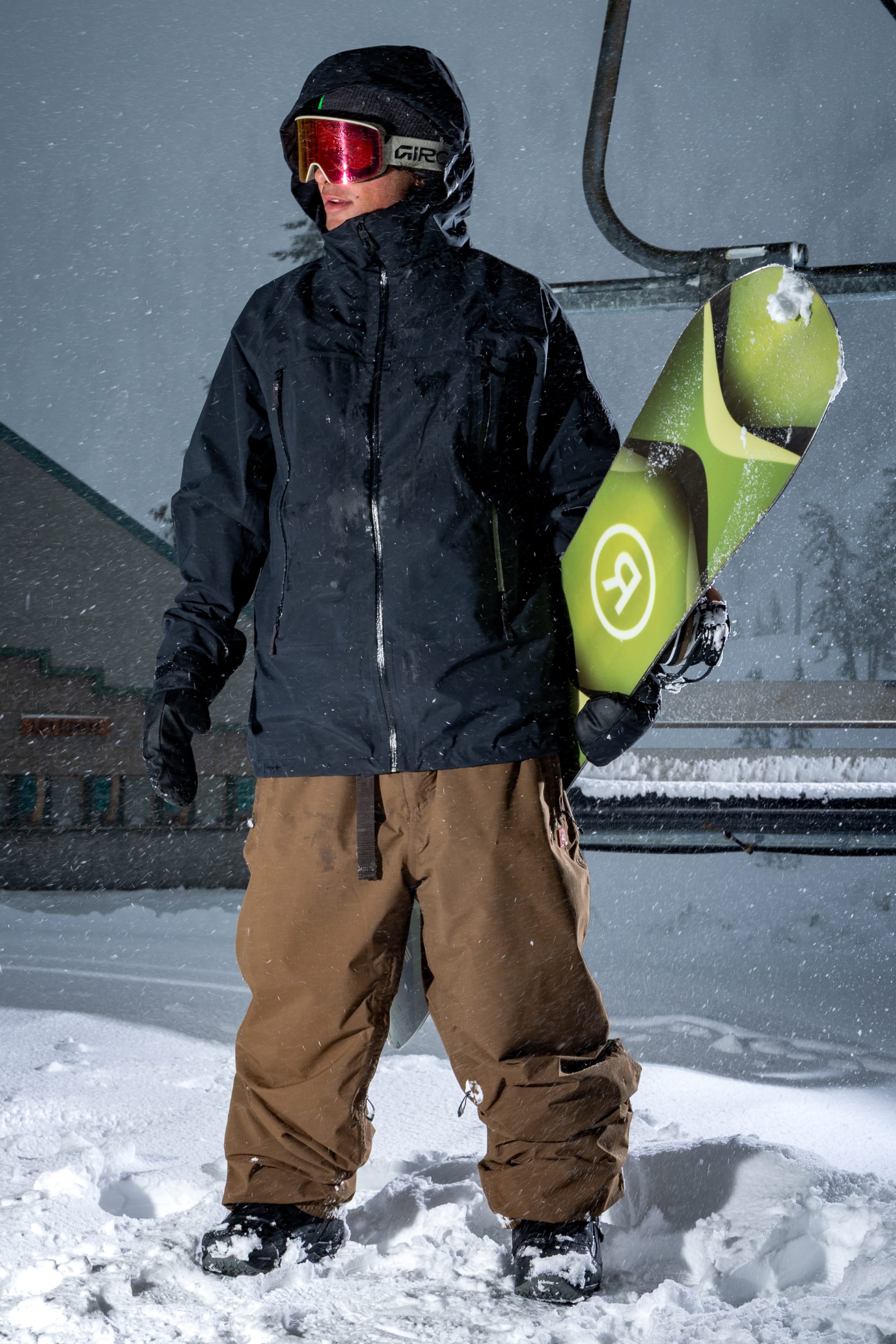 A snowboarder wearing the 686 Men’s SMARTY® 3-In-1 Rodeo™ 3L Jacket stands in the snow, dressed in brown pants and goggles and holding a snowboard with a green design. Snow is falling, and a ski lift can be seen in the background.