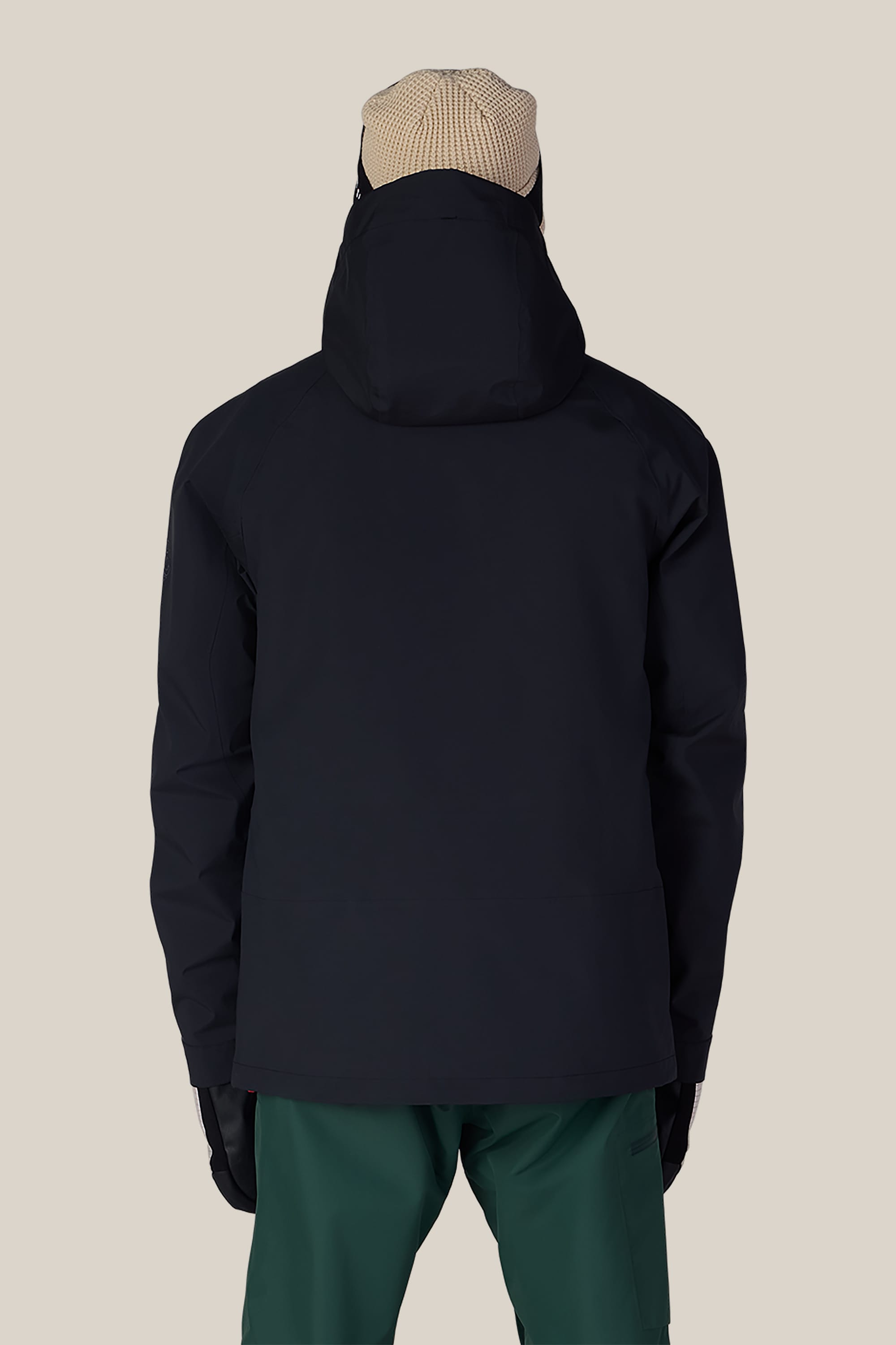 A person stands with their back to the camera, wearing the 686 Men’s SMARTY® 3-In-1 Rodeo™ 3L Jacket by 686, paired with green pants, black gloves, and a beige knit beanie against a plain light background.