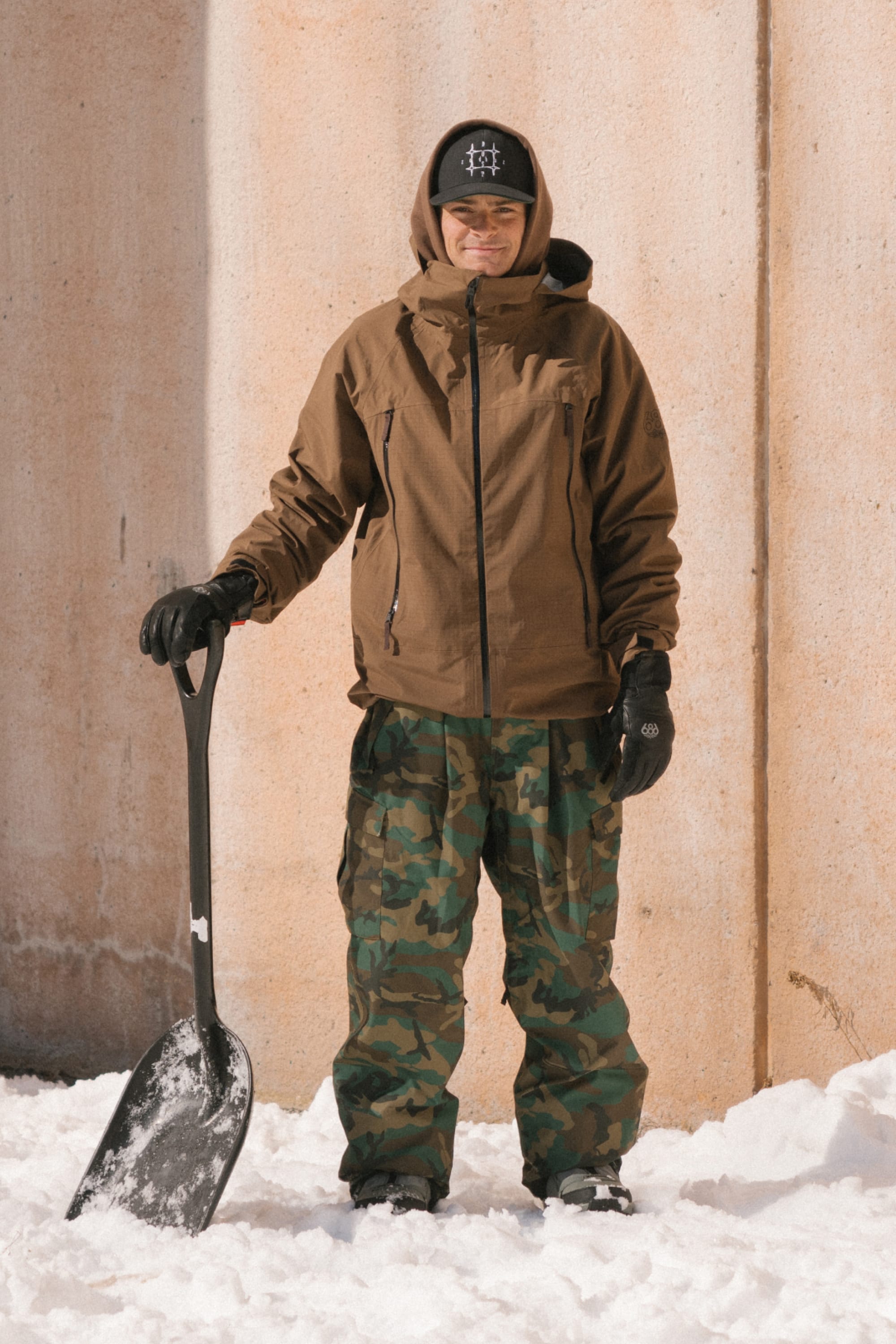 Wearing the 686 Men’s SMARTY® 3-In-1 Rodeo™ 3L Jacket, a person stands on snow in camo pants and black gloves, holding a snow shovel upright. They have on a black hat and are positioned in front of a tall concrete wall.