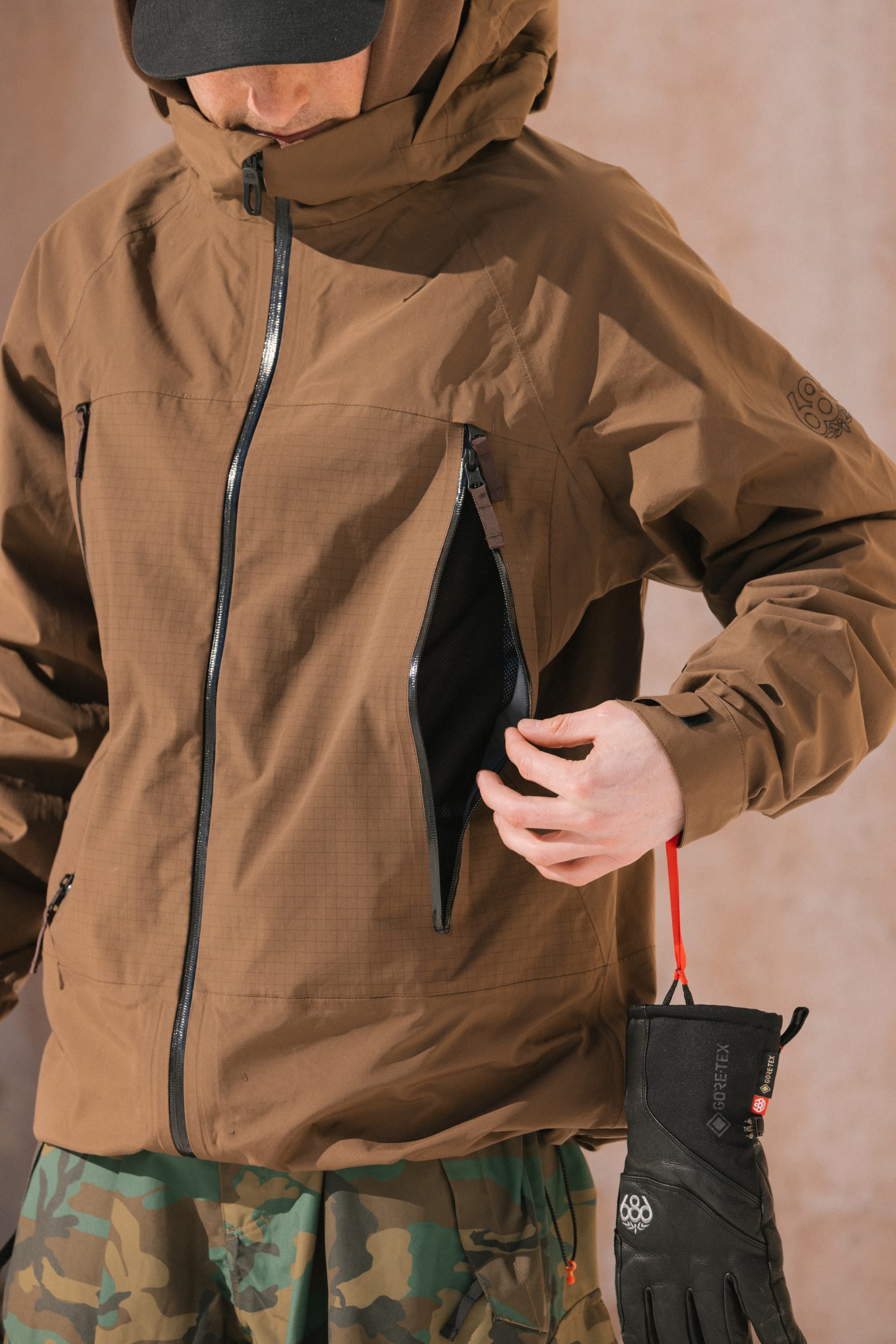 Wearing the 686 Men’s SMARTY® 3-In-1 Rodeo™ 3L Jacket in brown, a person unzips the chest pocket. They hold black winter gloves with a red loop in their left hand, and their face is partially covered by a black cap and camo pants.