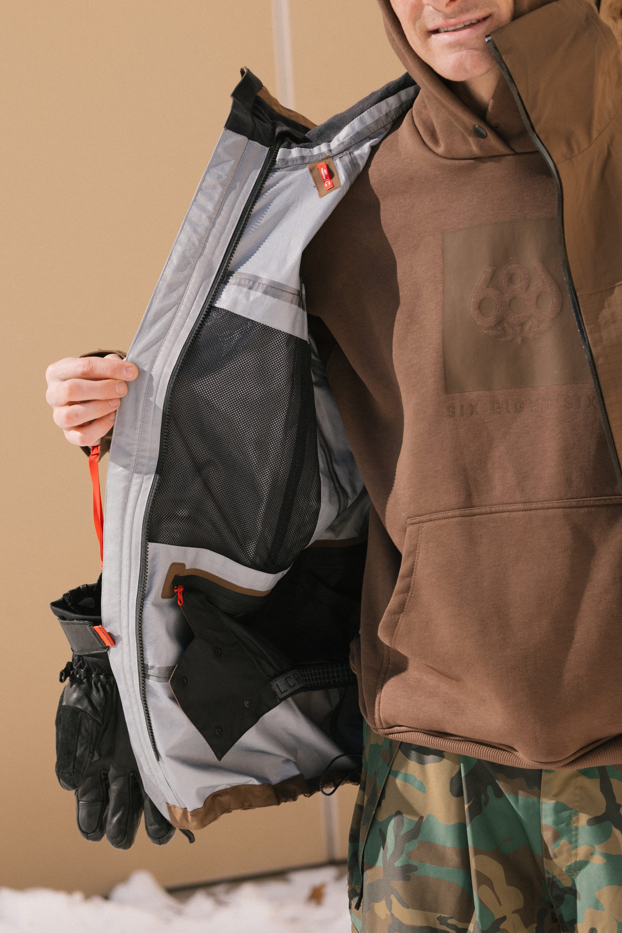 A person wearing the 686 Men’s SMARTY® 3-In-1 Rodeo™ 3L Jacket from 686 and camouflage pants shows off the jacket’s mesh and zippered interior pockets. Black gloves hang from their arm, and snow covers the ground.