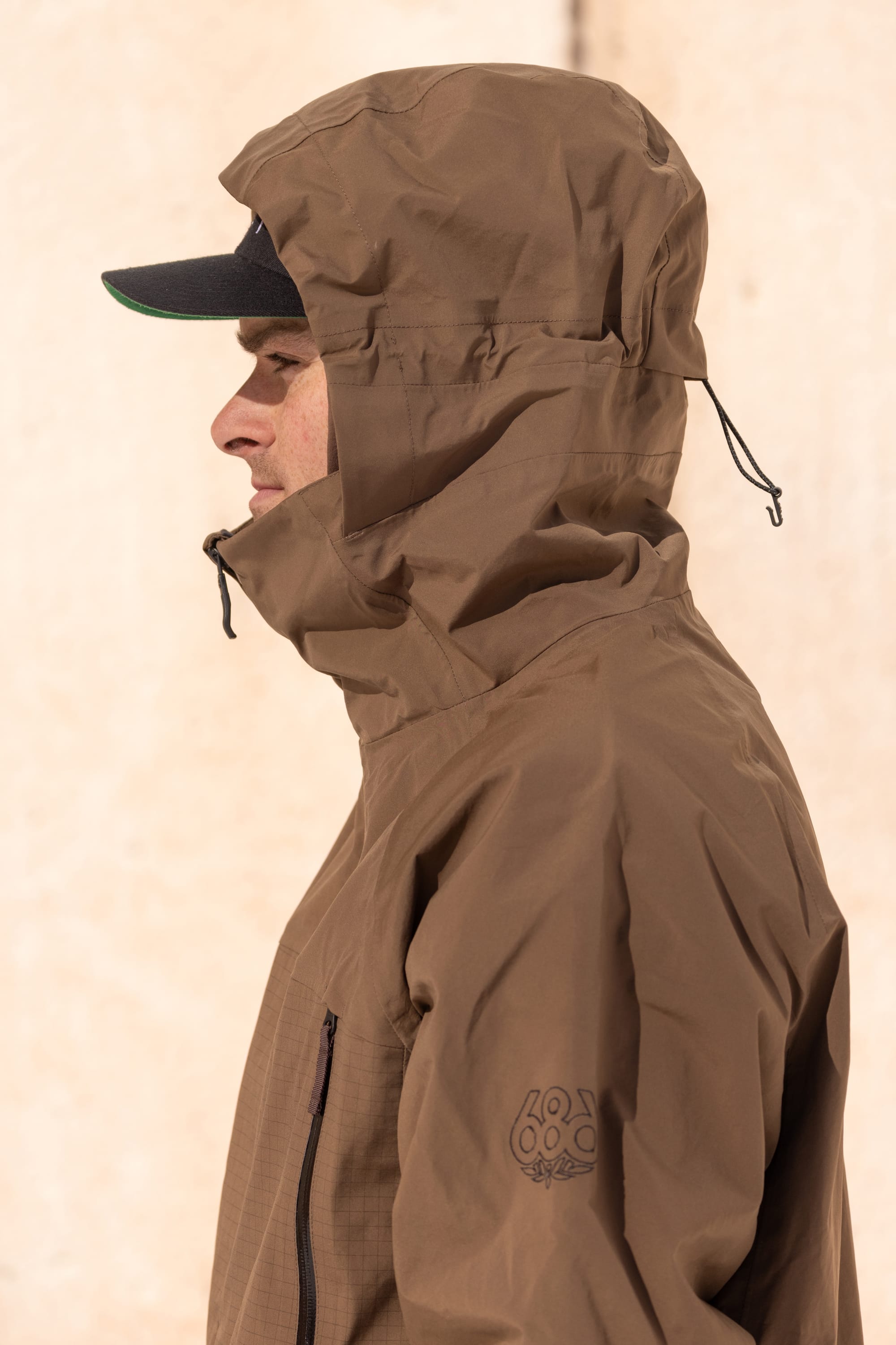 A person wearing the 686 Men’s SMARTY® 3-In-1 Rodeo™ 3L Jacket in brown and a black cap, shown in profile view, stands against a light beige background.