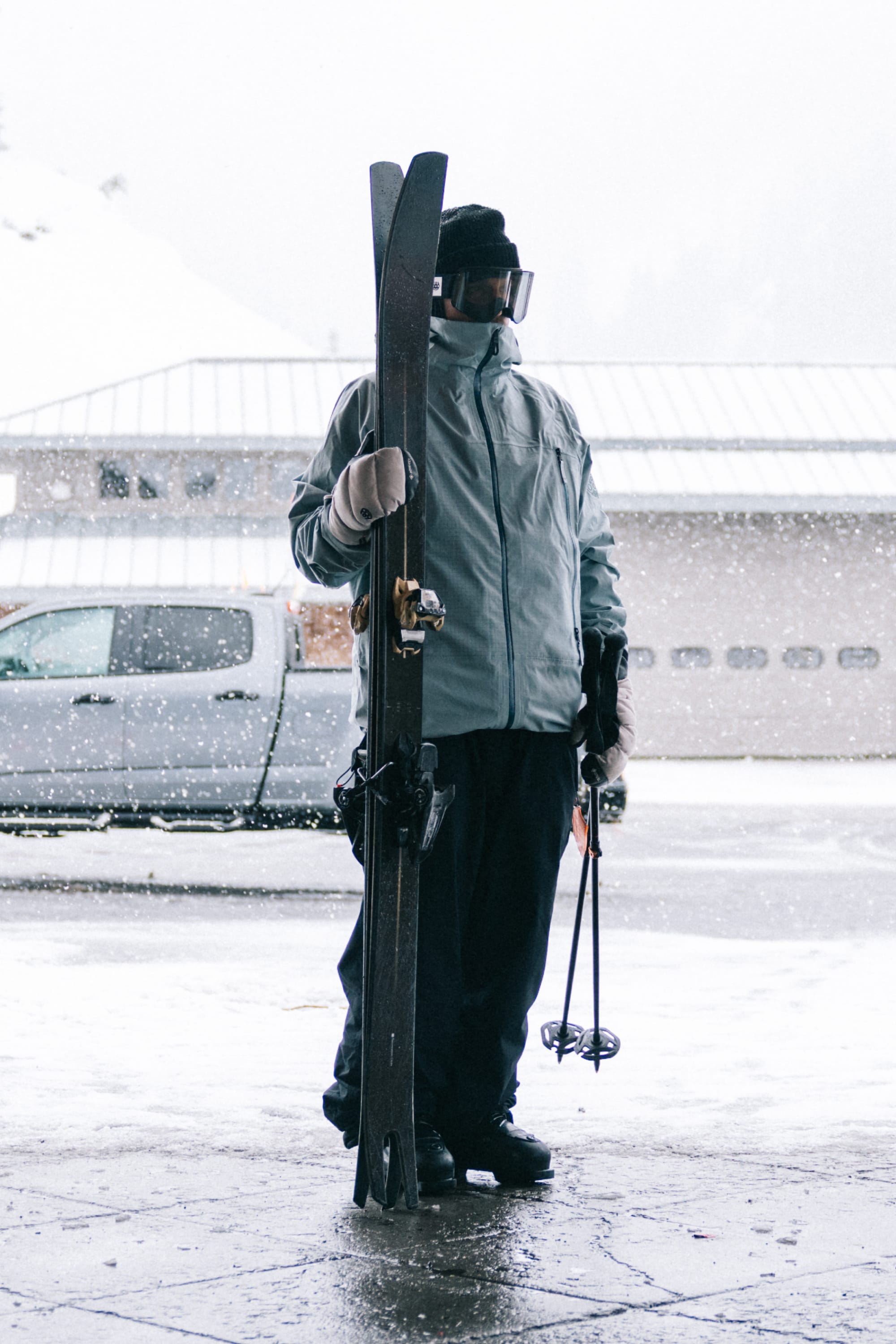 Wearing the 686 Men’s SMARTY® 3-In-1 Rodeo™ 3L Jacket by 686, a person stands outside in snowy weather near a parked vehicle and building, holding skis and poles.