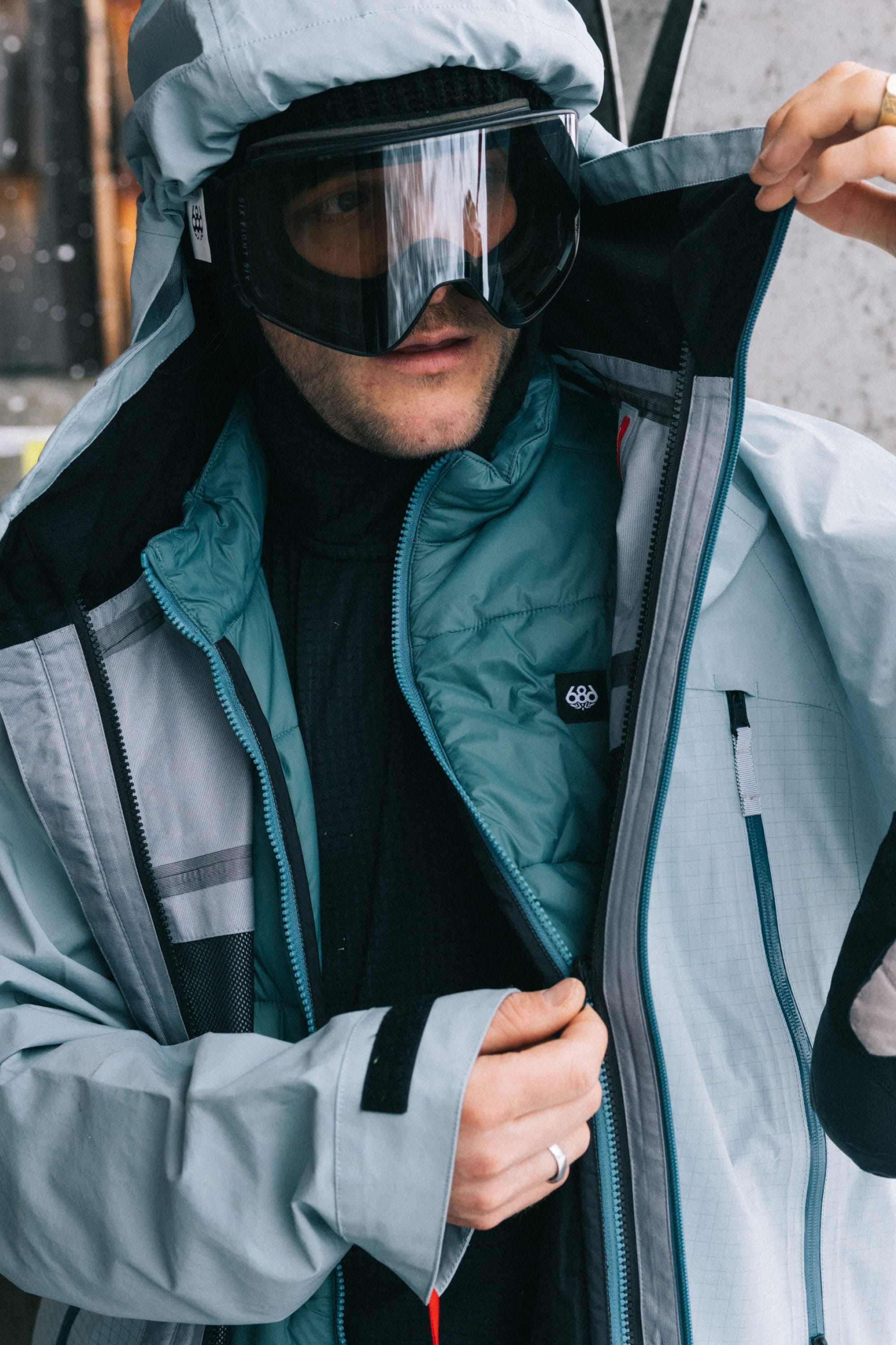 A person wearing the 686 Men’s SMARTY® 3-In-1 Rodeo™ 3L Jacket, along with a teal vest, black gloves, and ski goggles, adjusts their coat while standing outside near a concrete wall.