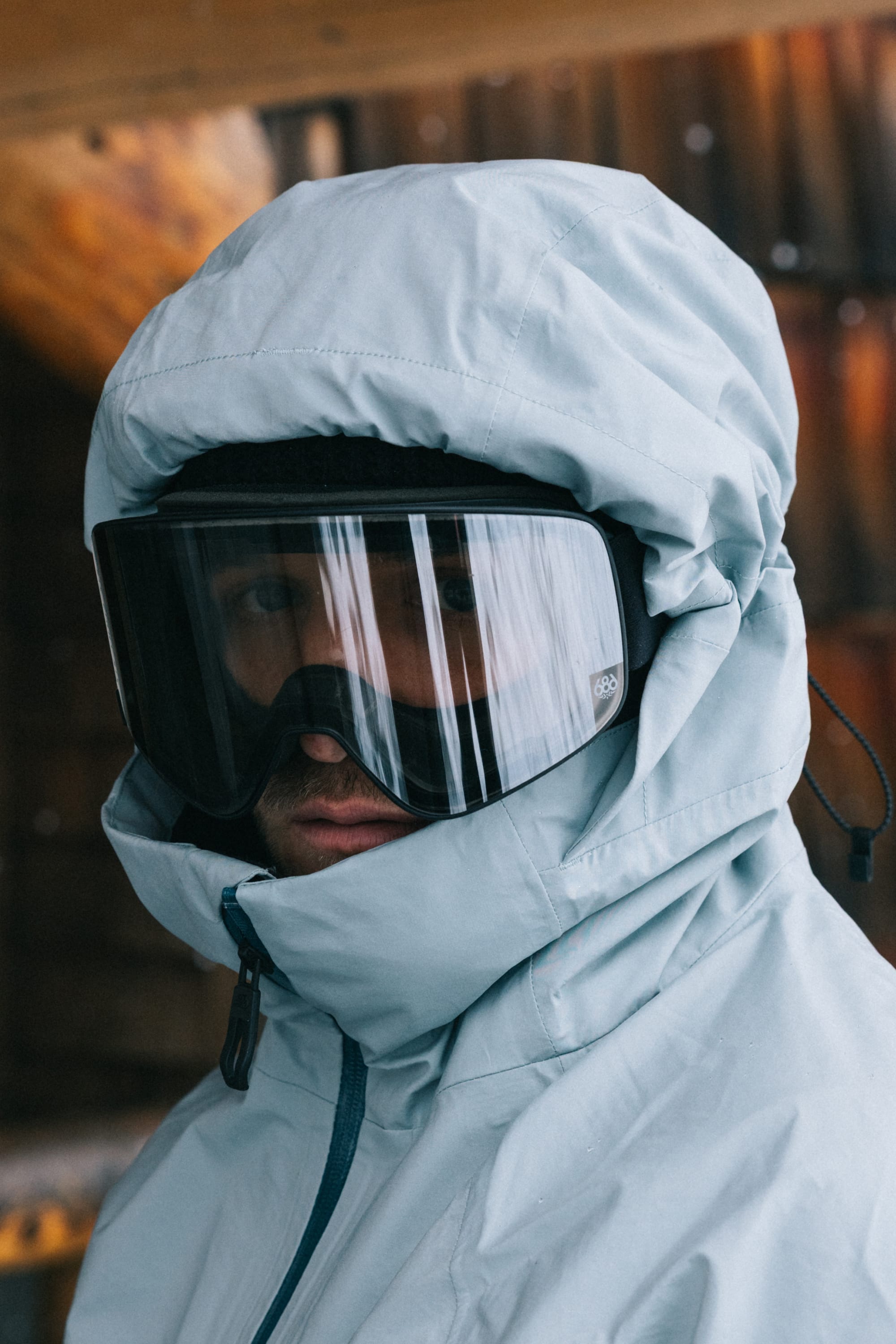 A person wearing the 686 Men’s SMARTY® 3-In-1 Rodeo™ 3L Jacket in light blue and black ski goggles stands outside near a wooden wall, looking toward the camera.