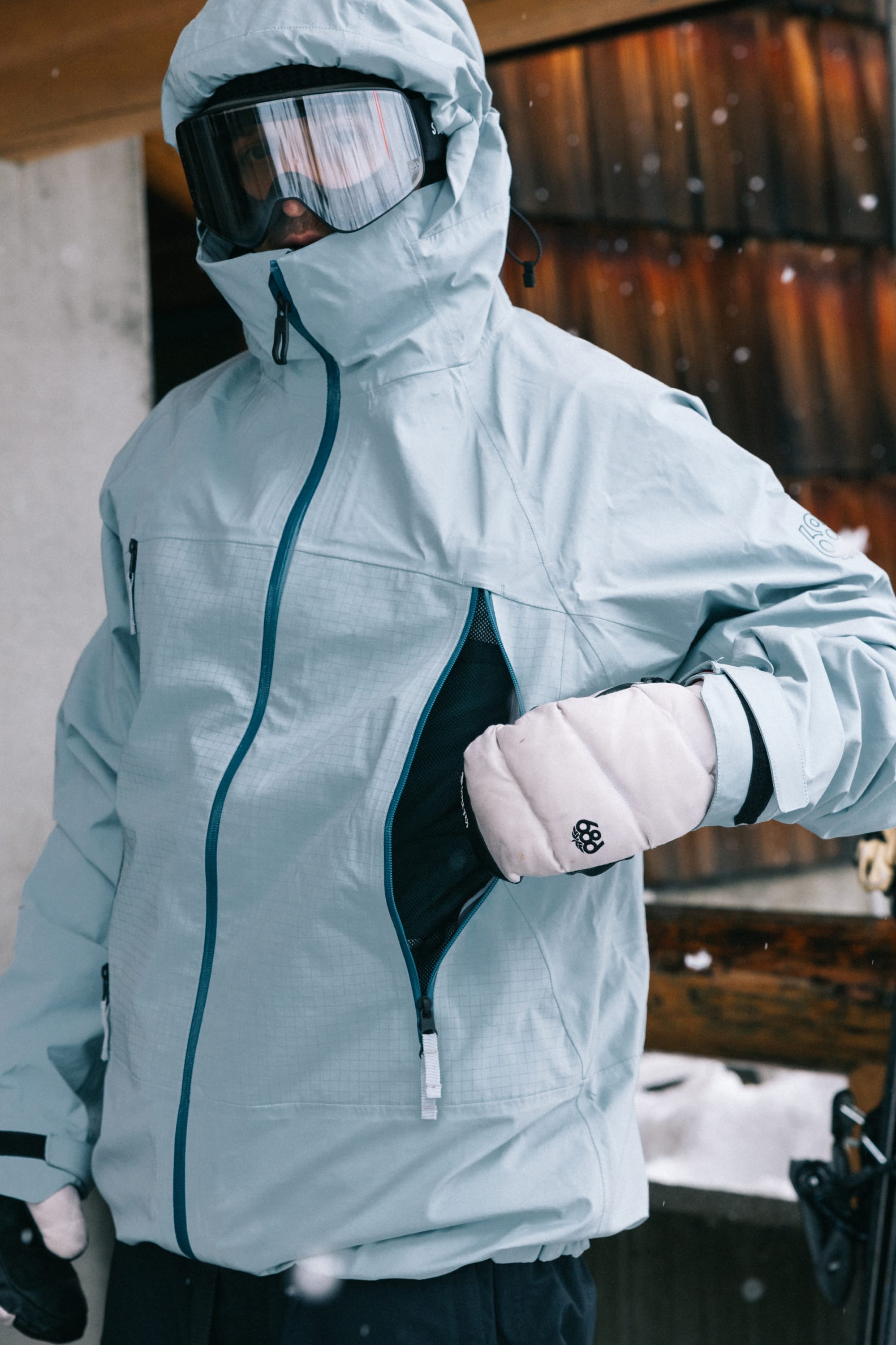 A person outdoors in snowy weather, unzipping a side pocket of the 686 Men’s SMARTY® 3-In-1 Rodeo™ 3L Jacket, paired with ski goggles and white mittens.