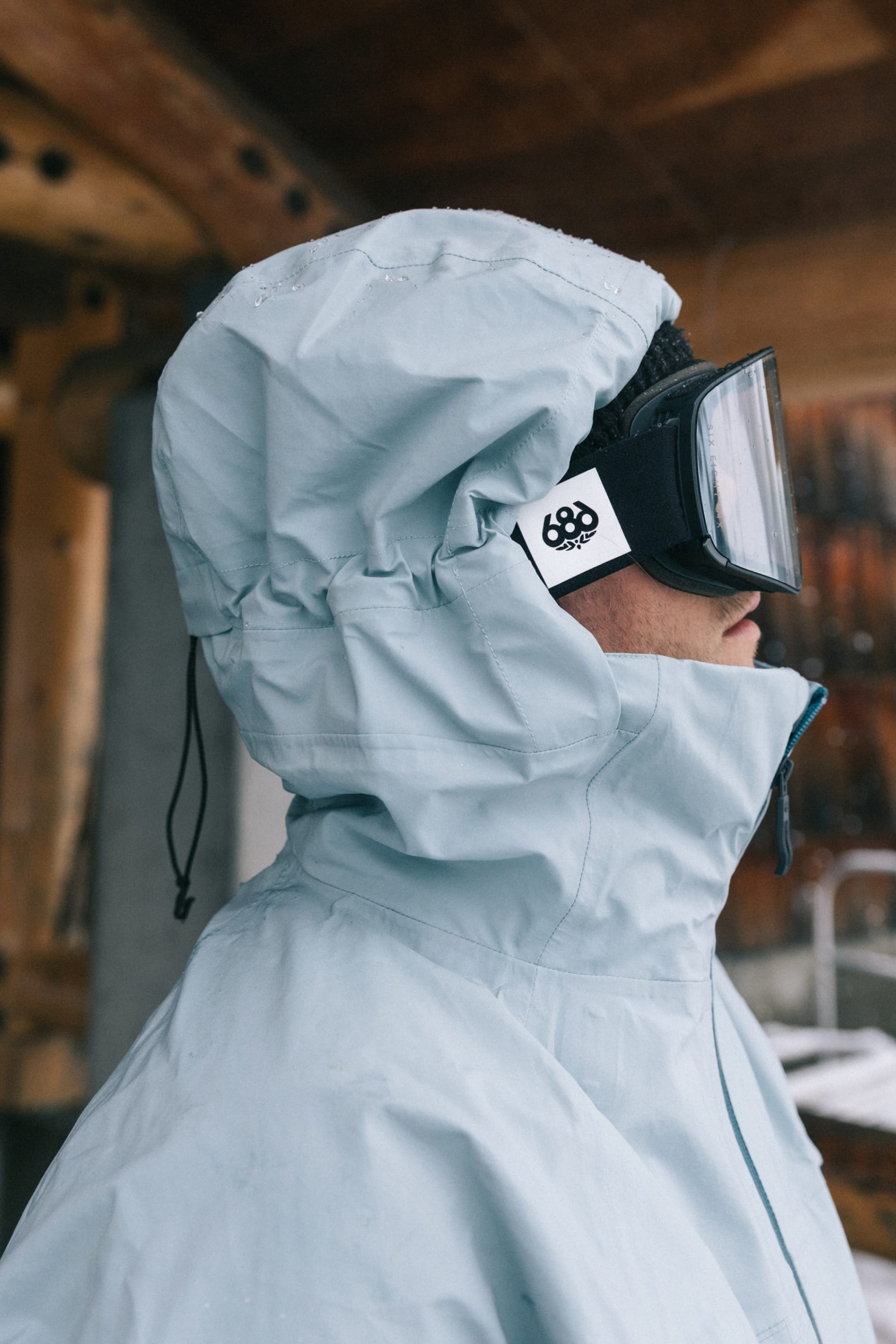 A person wearing the 686 Men’s SMARTY® 3-In-1 Rodeo™ 3L Jacket in light blue, along with ski goggles, stands outdoors in a snowy setting facing left. Wooden beams and a blurred wintry landscape are visible in the background.