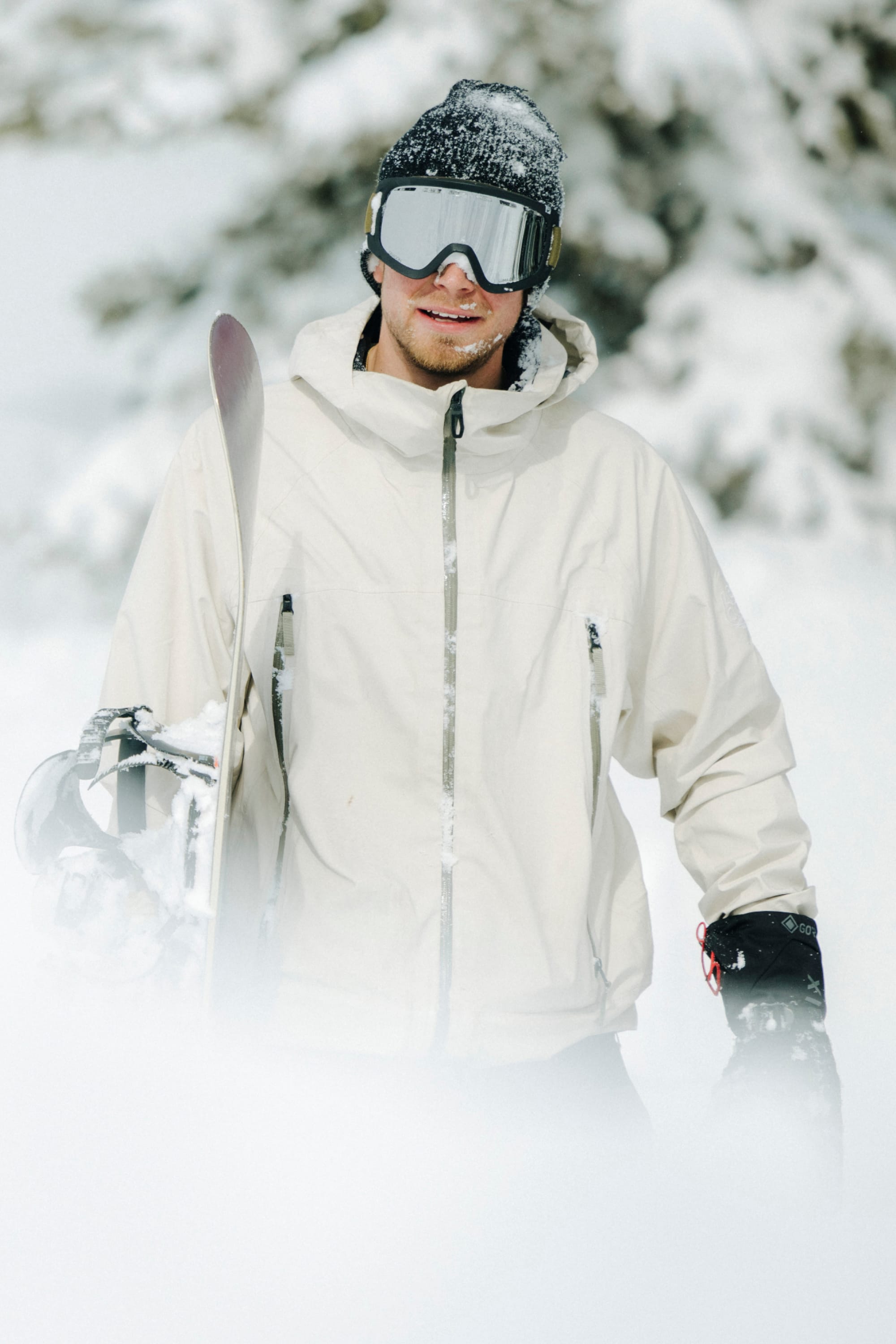 Clad in the 686 Men’s SMARTY® 3-In-1 Rodeo™ 3L Jacket, a person stands amid snowy terrain, wearing black gloves and goggles and holding skis, with snow blanketing the ground and trees behind them.