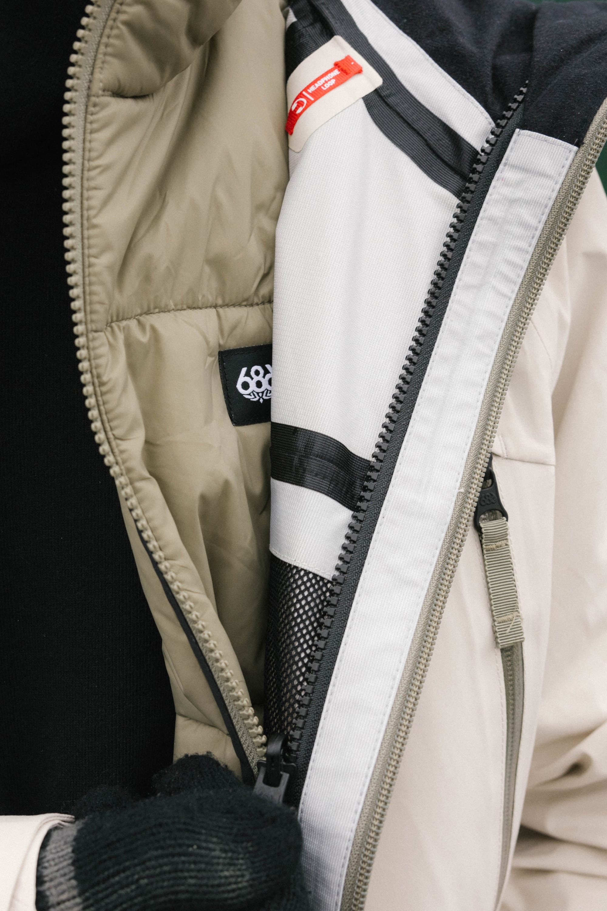 A close-up of a person unzipping the 686 Men’s SMARTY® 3-In-1 Rodeo™ 3L Jacket, revealing inner layers and black gloves. Visible tags include the 686 brand logo and a red label.