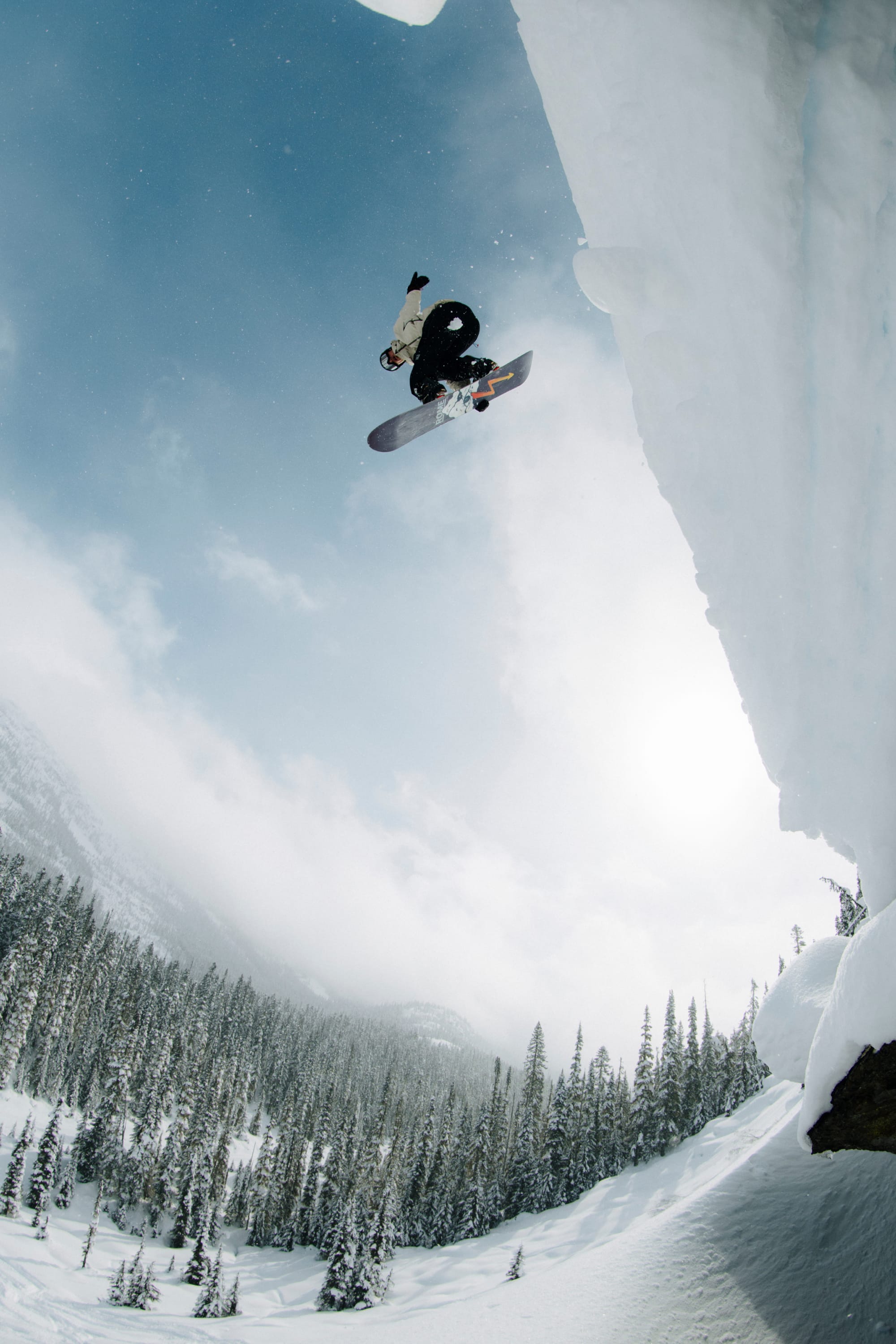 Wearing the 686 Men’s SMARTY® 3-In-1 Rodeo™ 3L Jacket by 686, a snowboarder performs a mid-air trick off a snowy cliff, framed by snow-covered trees and mountains beneath a cloudy sky.
