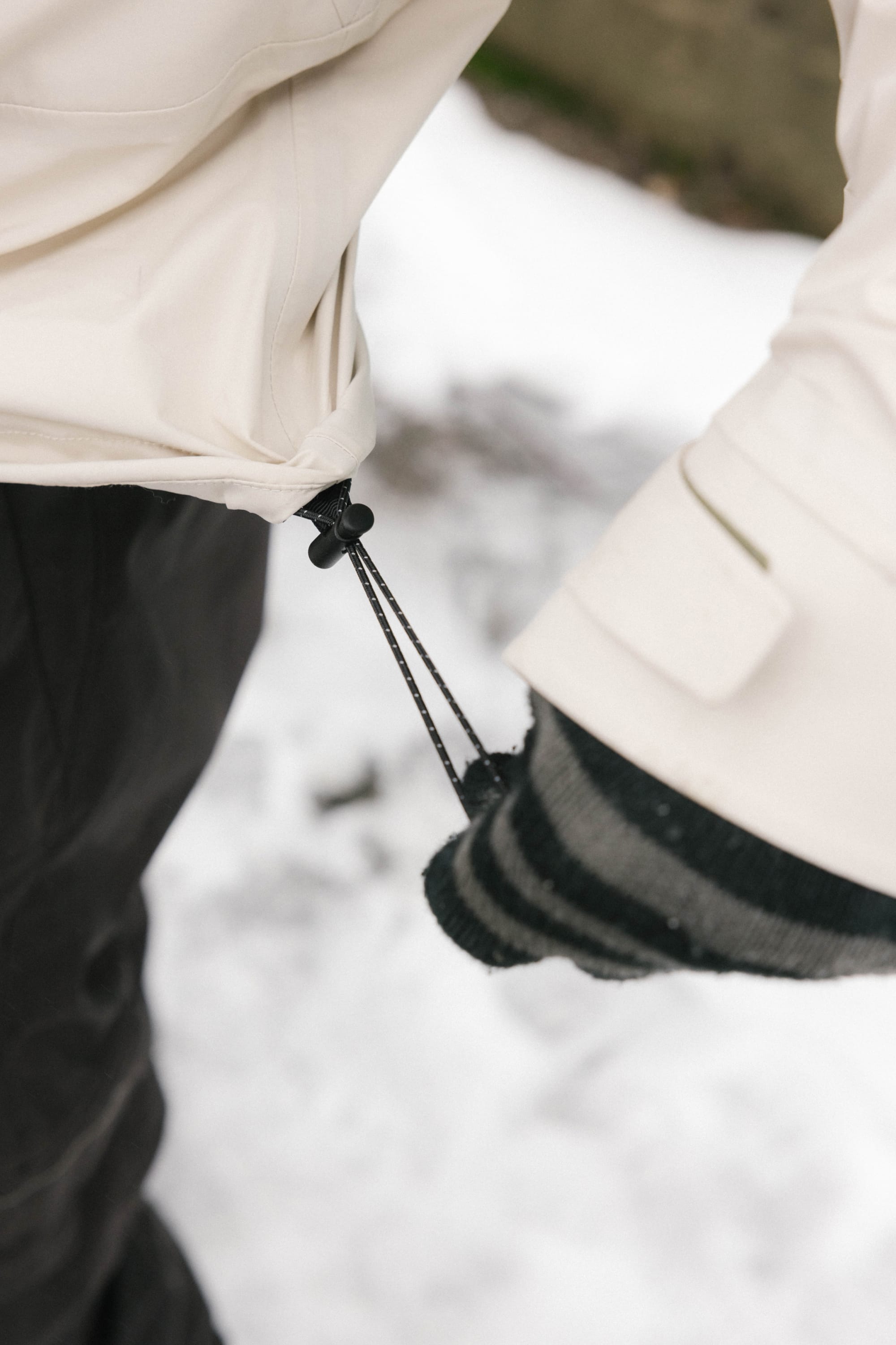 A person wearing black and gray striped gloves tightens the drawstring on a 686 Men’s SMARTY® 3-In-1 Rodeo™ 3L Jacket while standing on snow.