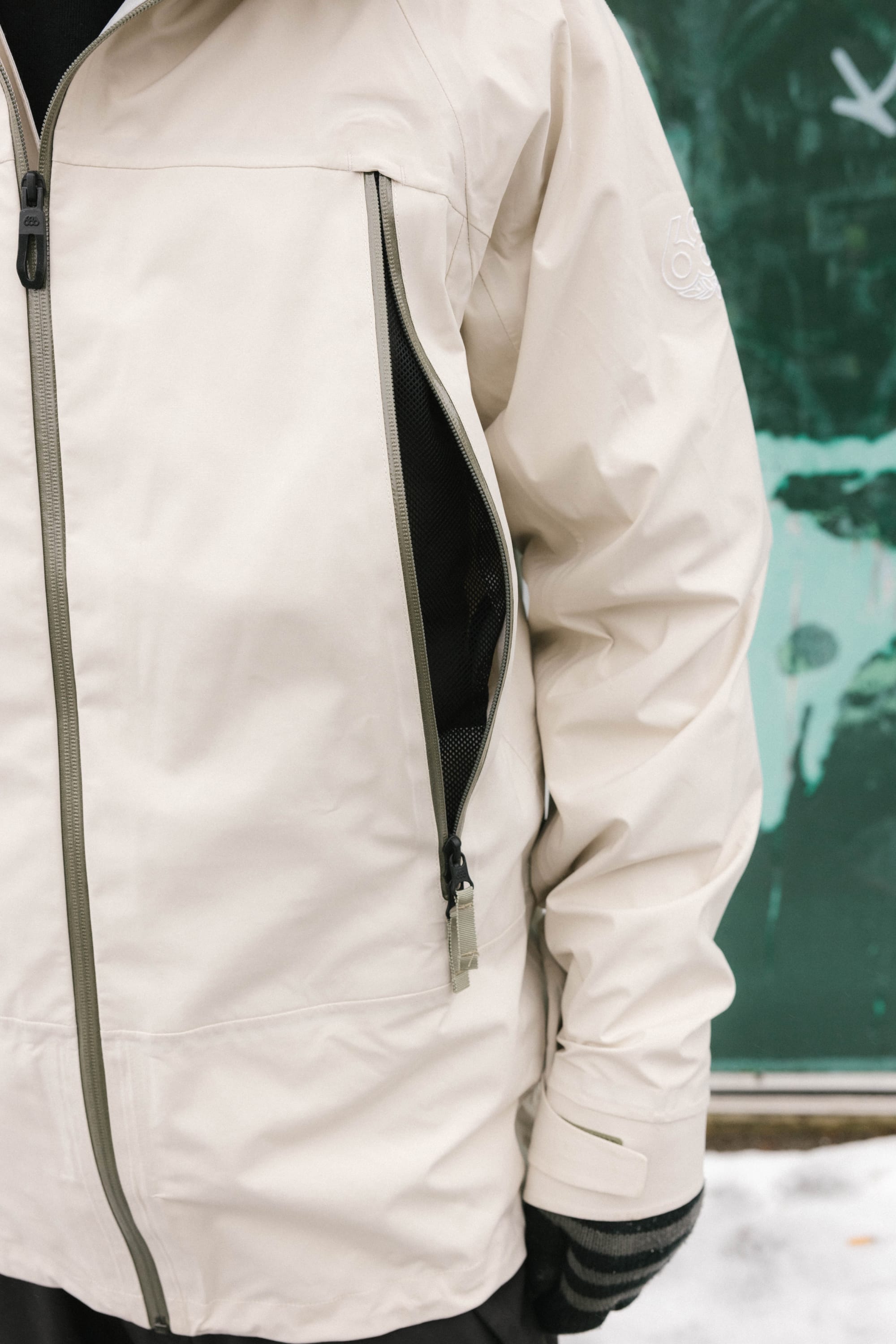 A person wearing the 686 Men’s SMARTY® 3-In-1 Rodeo™ 3L Jacket, which features a vertical zippered pocket and a knit cuff on the left sleeve, stands in front of a green wall with white graffiti.
