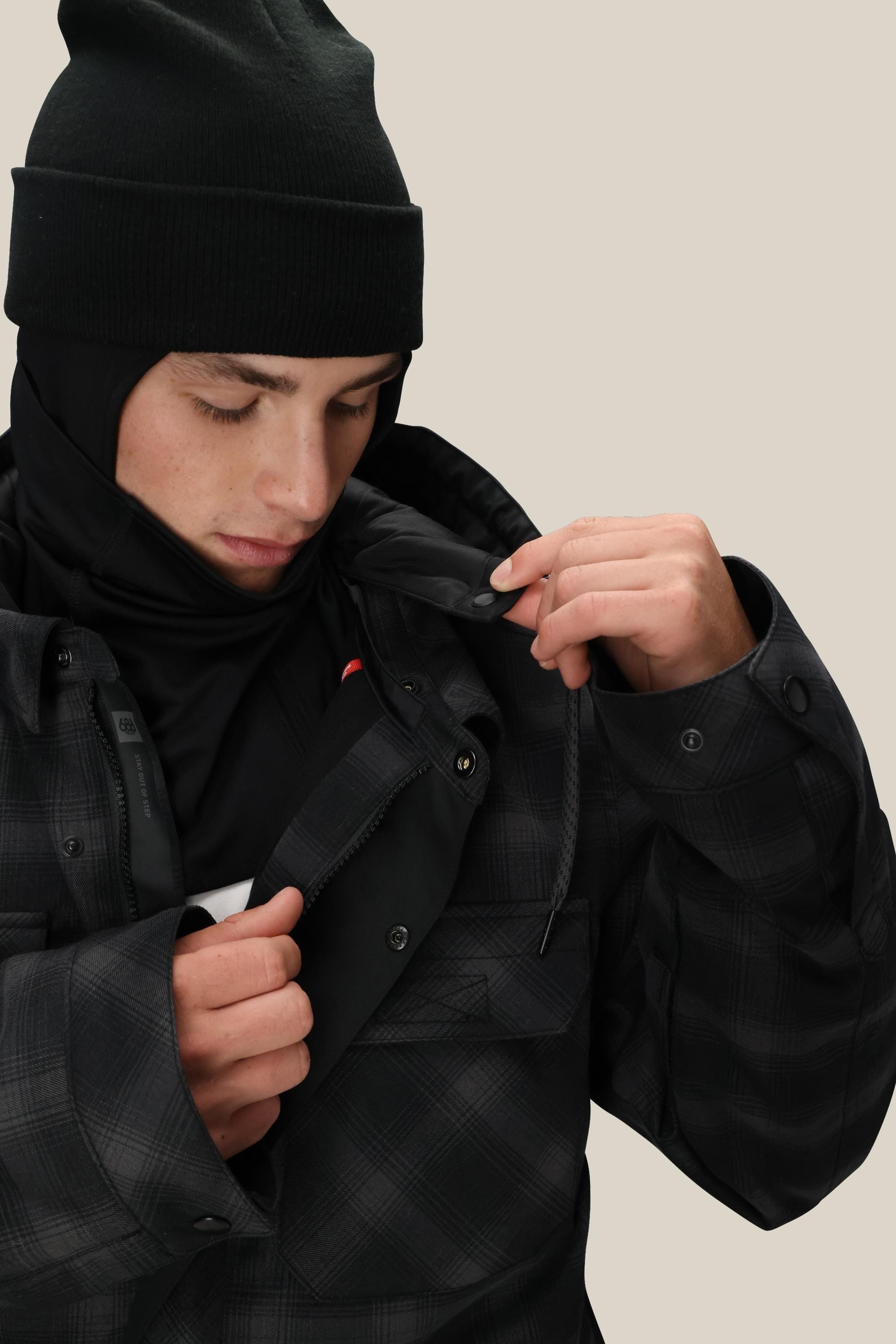 A person wearing a black beanie, black base layer, and the 686 Mens Woodland Insulated Jacket by 686 looks down while unzipping the jacket against a plain beige background.