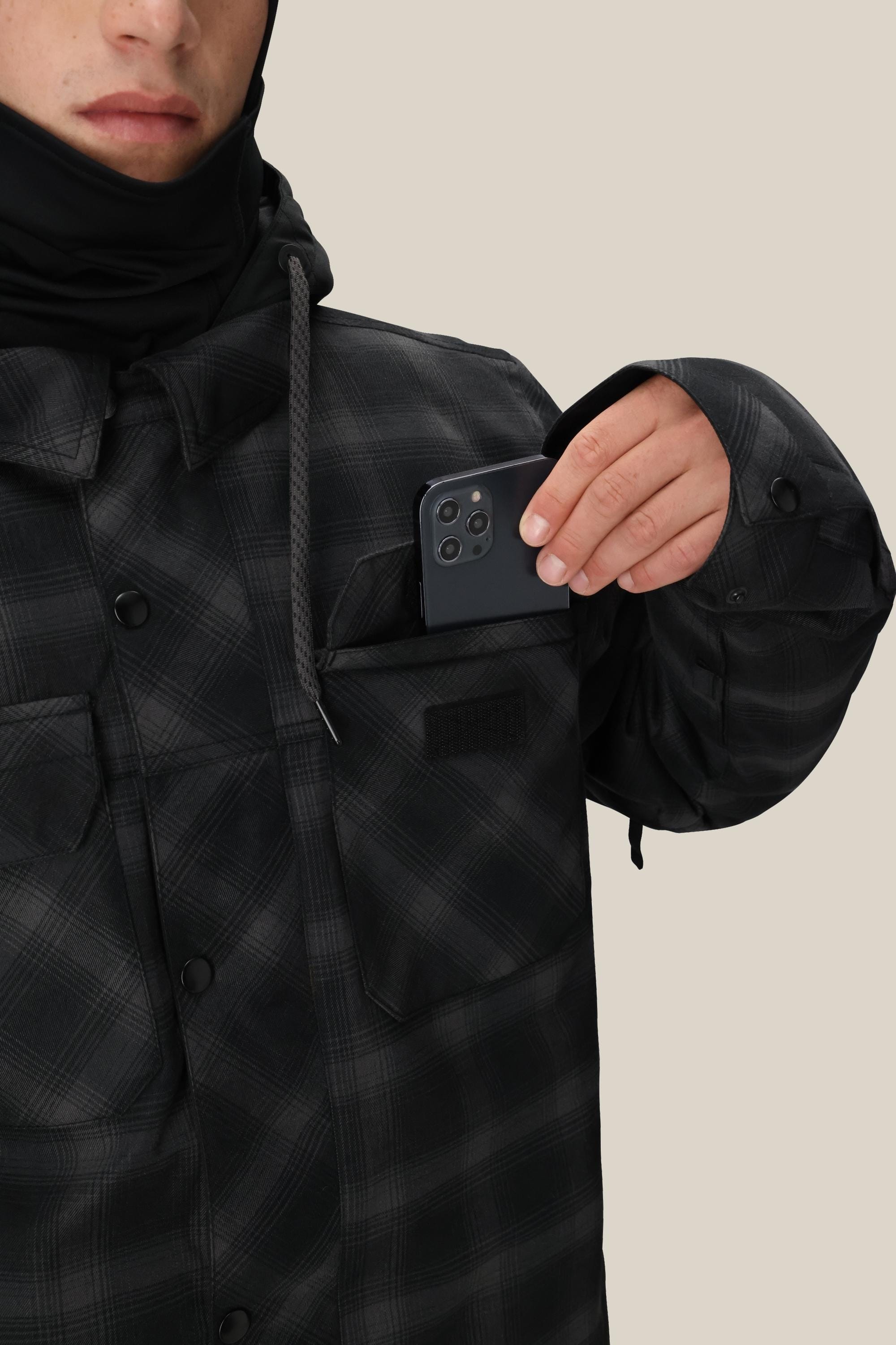 A person in a 686 Mens Woodland Insulated Jacket is slipping a black smartphone with three cameras into their chest pocket. Their face is partly visible against a plain beige background.