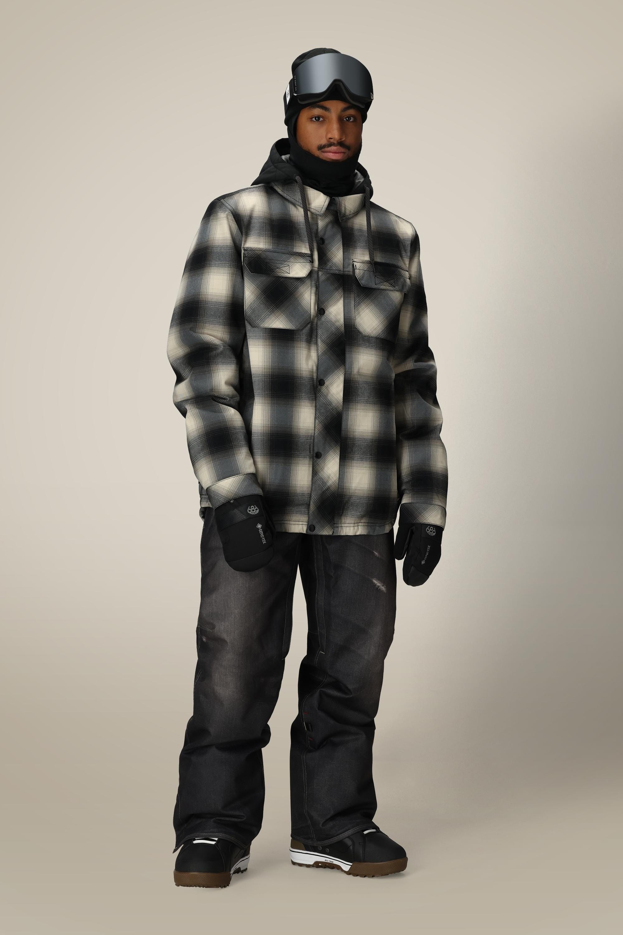 A man wearing the 686 Mens Woodland Insulated Jacket in black and white plaid from 686, paired with black snow pants, gloves, beanie, and ski goggles, stands against a plain light background.